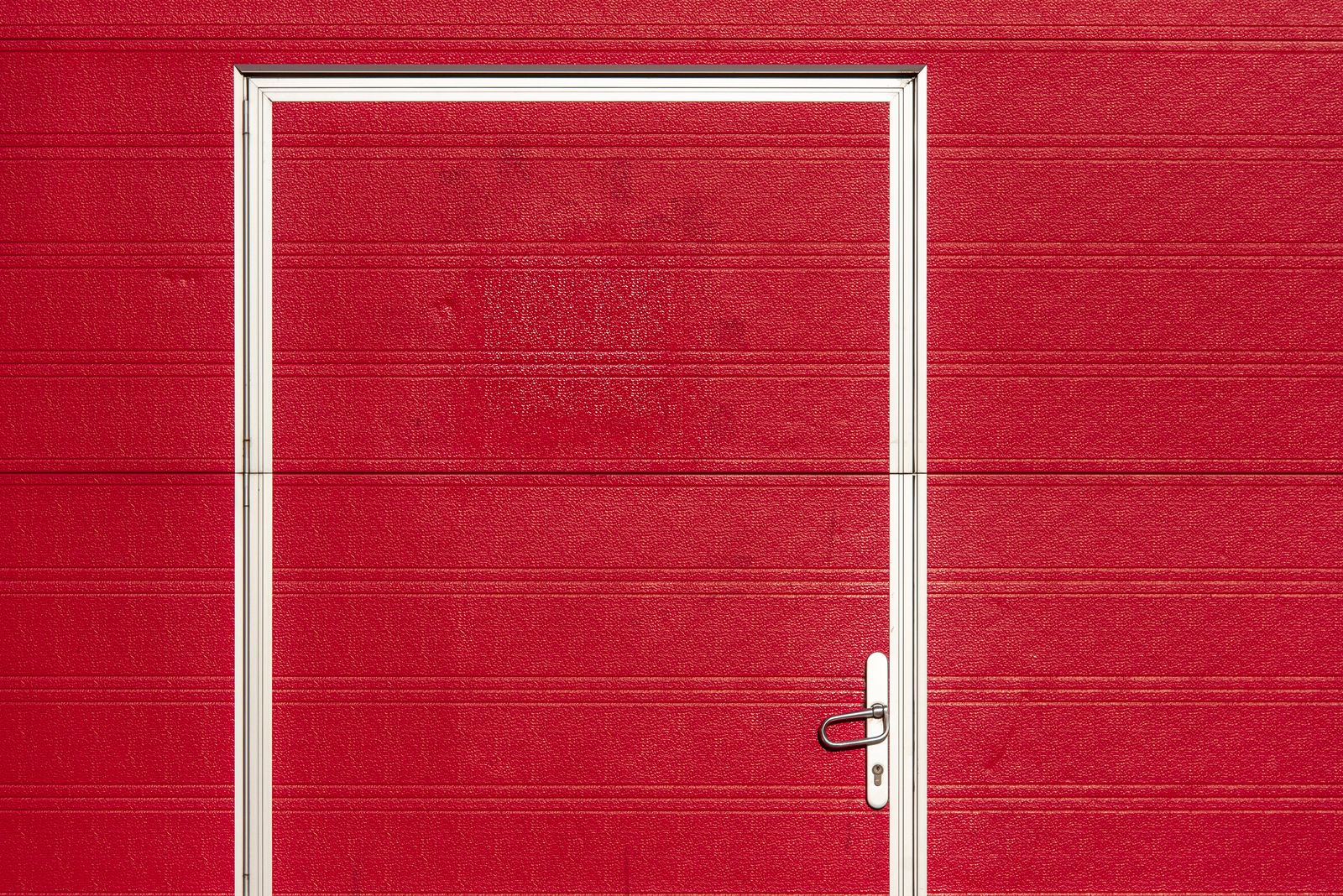 Close-up of a red textured sectional garage door with an integrated pass/wicket door framed in silver; a brushed-steel lever handle and keyhole sit on the lower right of the inset door.