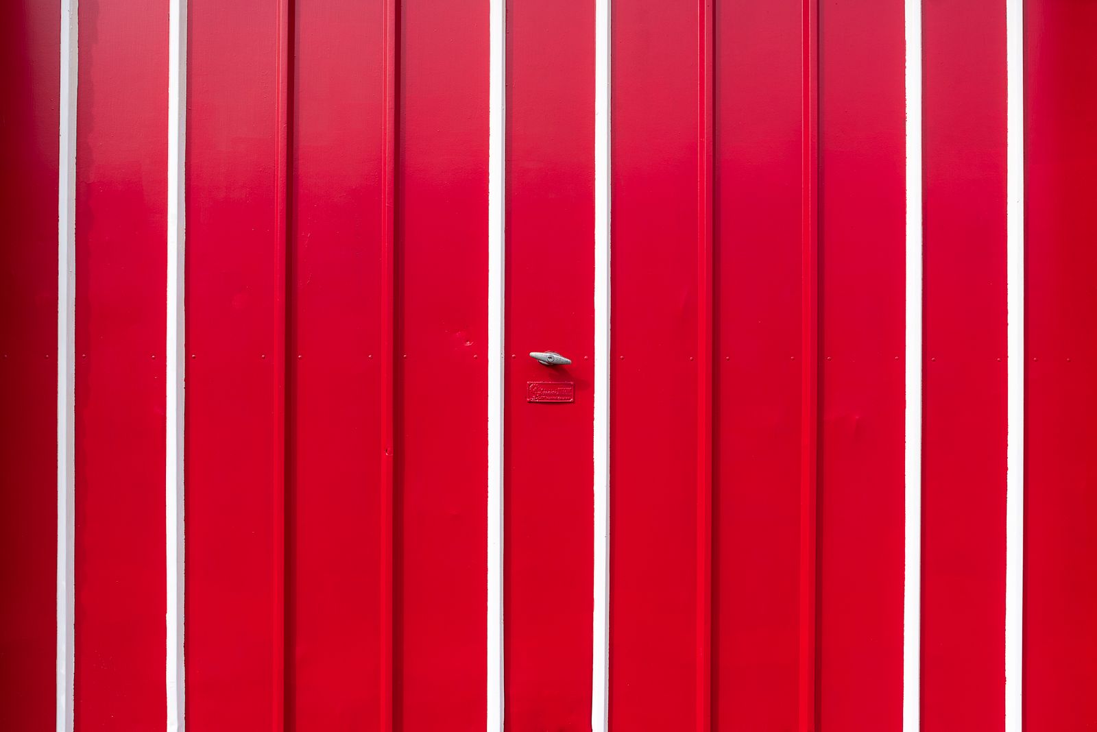 Tight shot of a glossy red ribbed metal garage door with white vertical seams; a small silver T-handle/latch and a red lock plate centered on the panel.