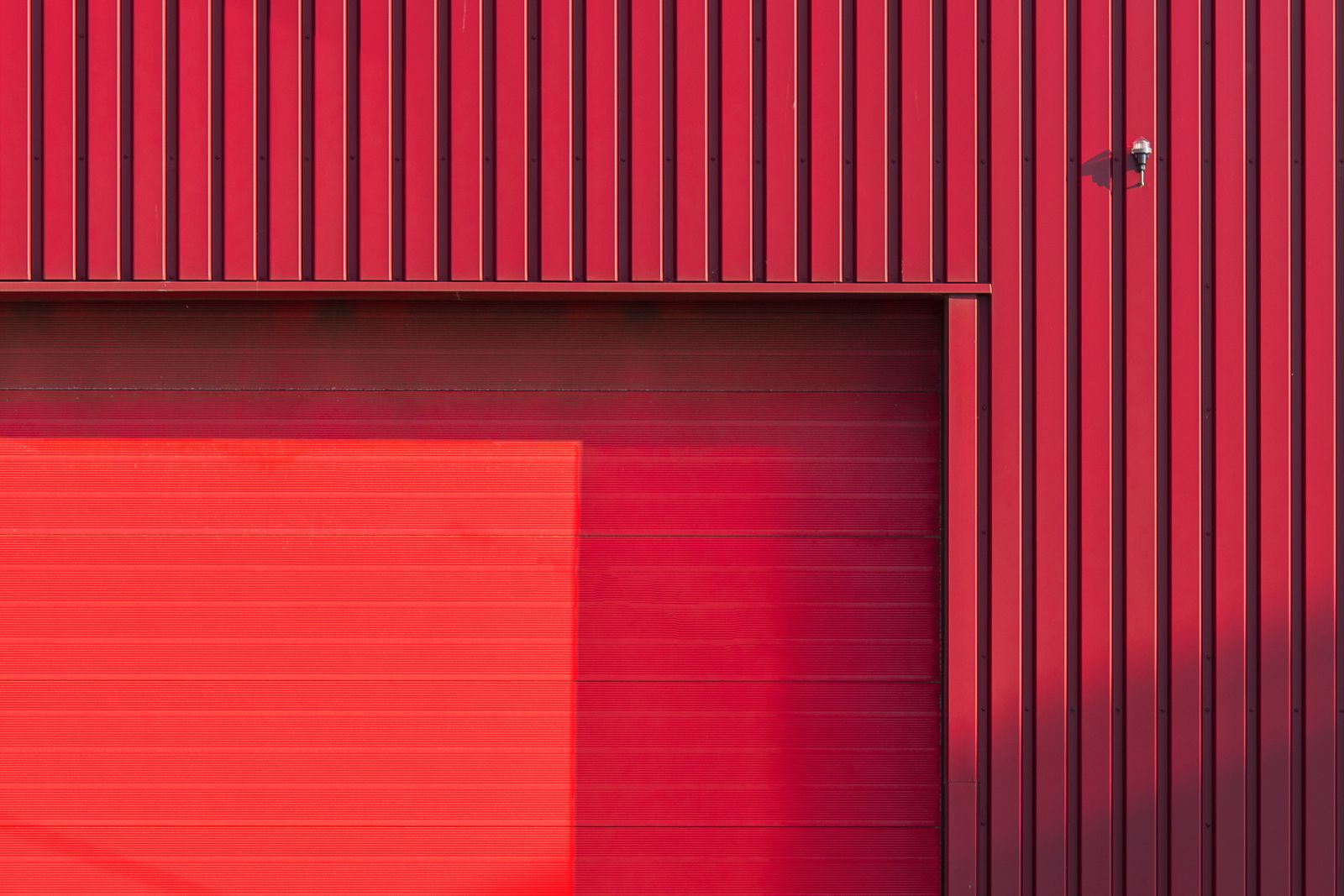 Corner detail of a deep-red ribbed sectional garage door meeting vertical red wall cladding; horizontal-ridge panel texture with strong directional light and a small wall fixture.