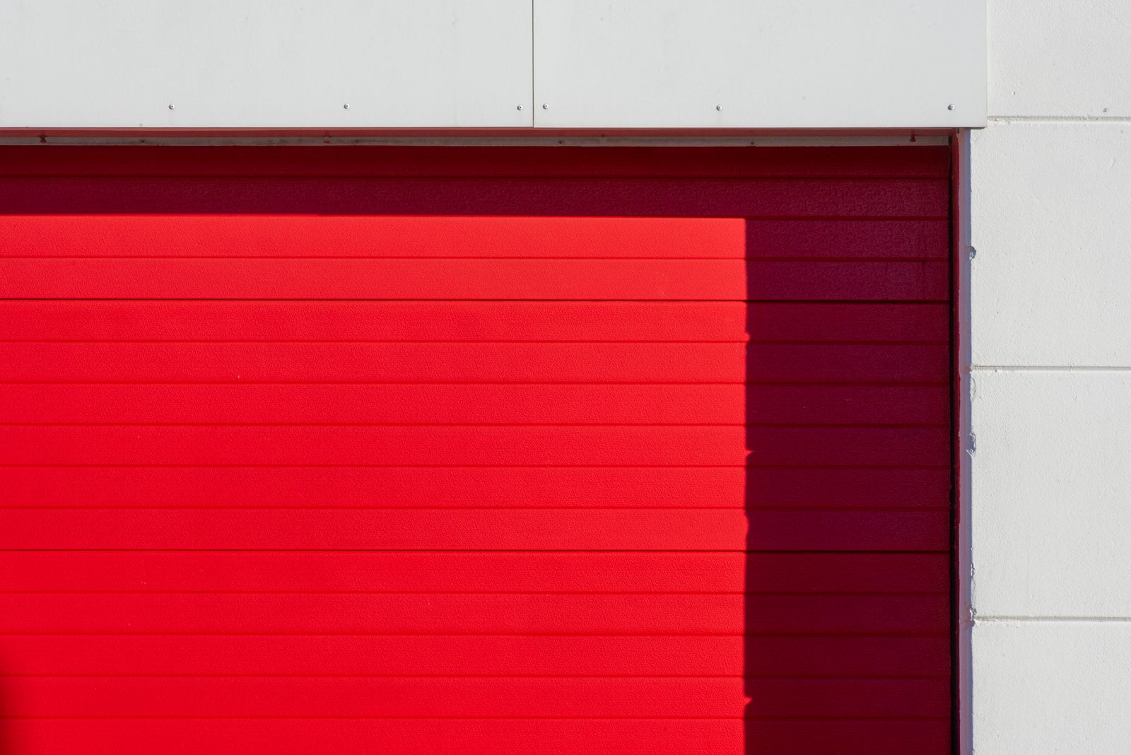 Tight diagonal-light shot of a bright-red slatted (sectional/roller) garage door panel with crisp horizontal ribs, set against white cladding and a concrete-block wall edge.