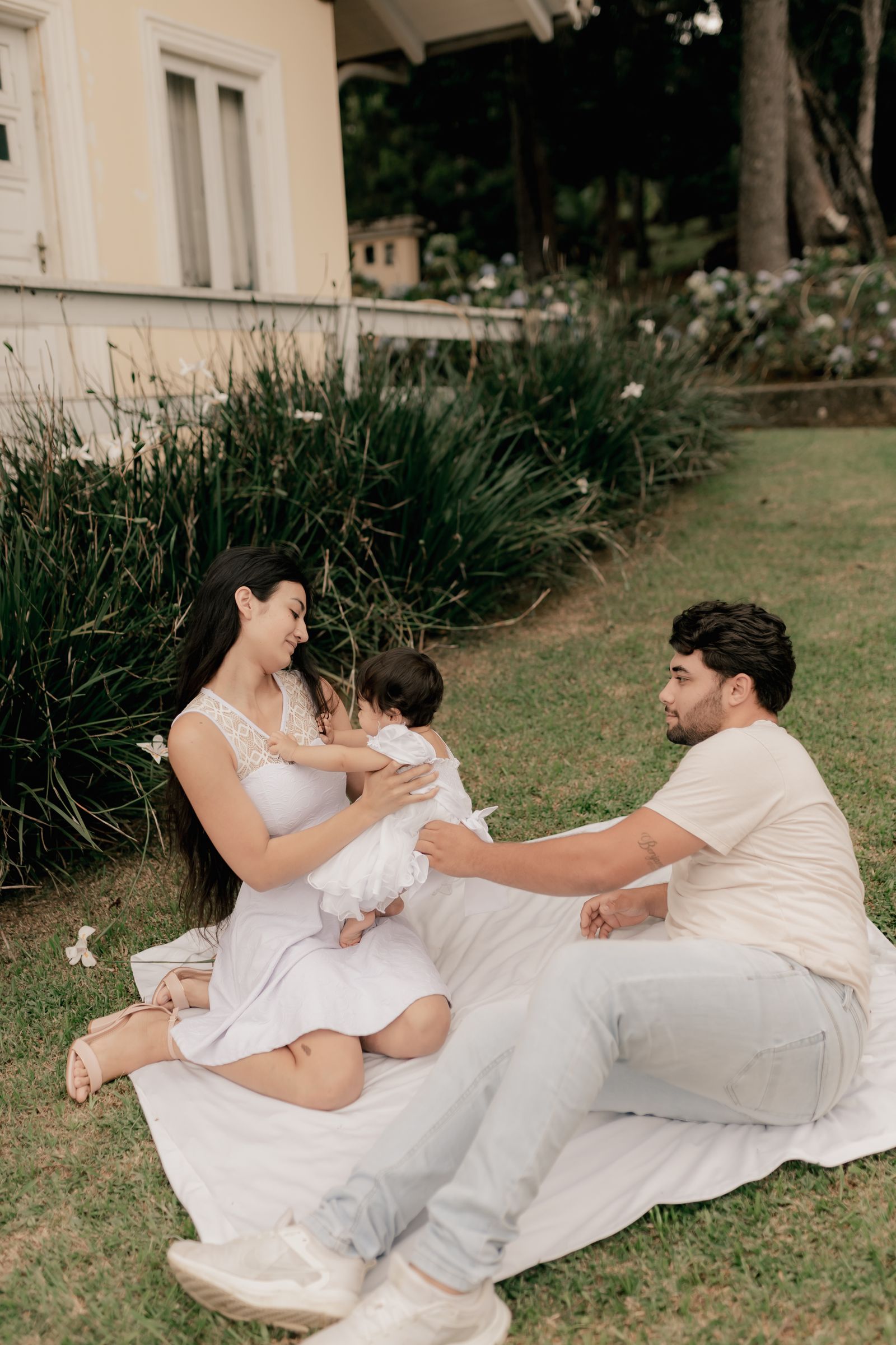 A young family (mom, dad, baby) relaxing on a white blanket on a grassy lawn beside a cozy light-colored house with white-flower garden beds; soft, airy natural light.