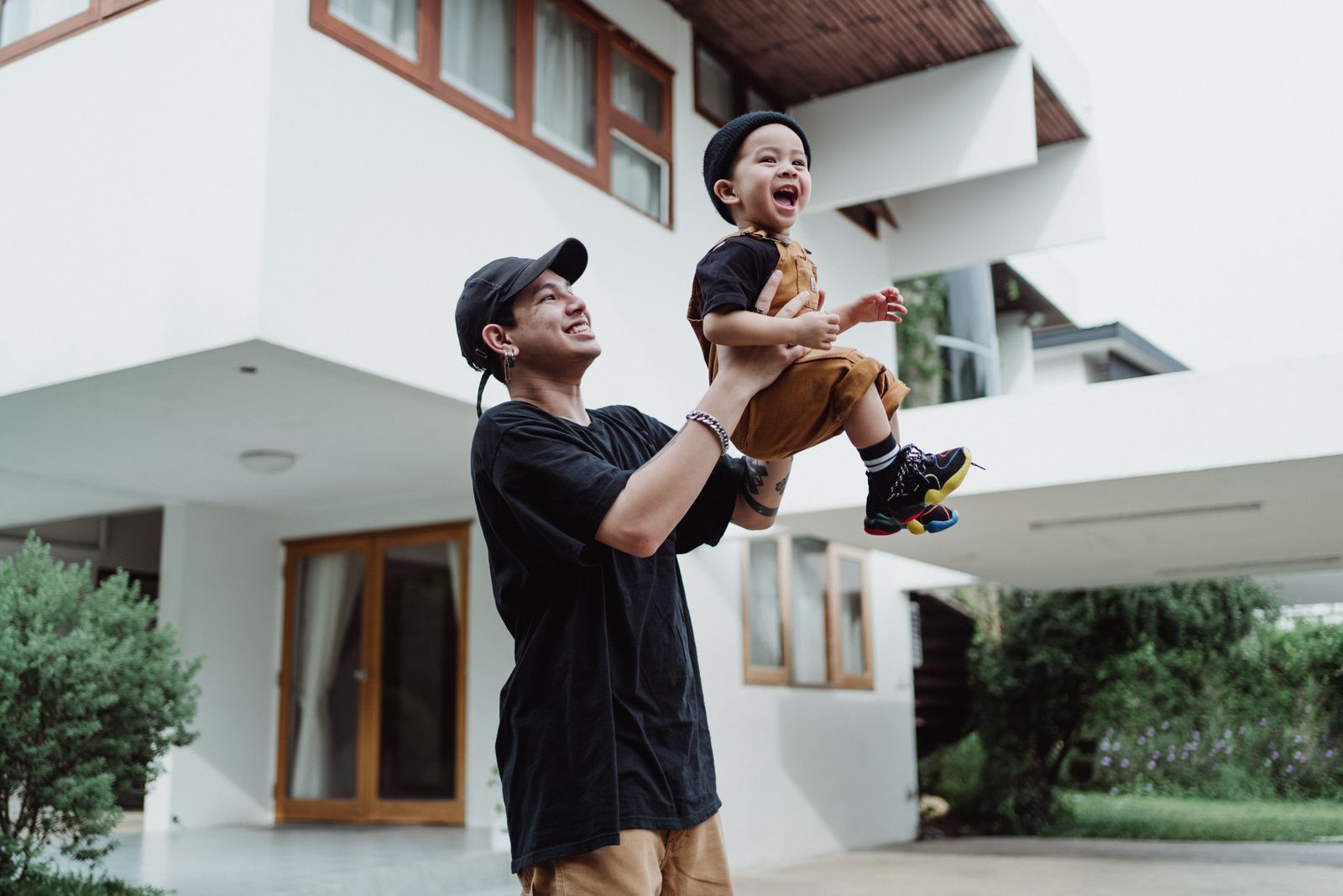 A smiling father in a black tee and cap lifting his laughing toddler up into the air on the driveway in front of a modern white two-story home with wood-trim windows.