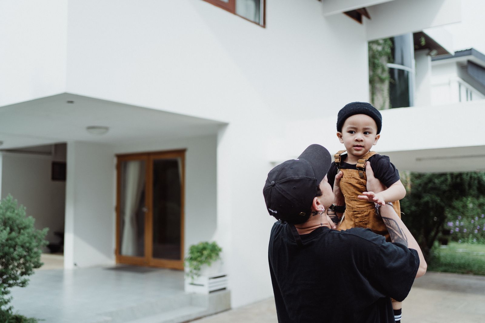 A father holding his toddler (in brown overalls) facing a modern white home with a wood double-door entry and concrete driveway; family arrival/home moment.