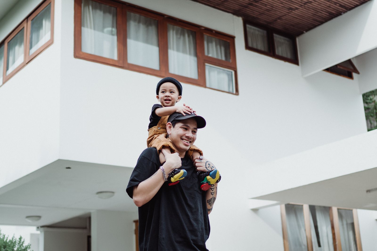 A father carrying a happy young child on his shoulders, both smiling, standing in front of a modern white home with wood-framed windows.