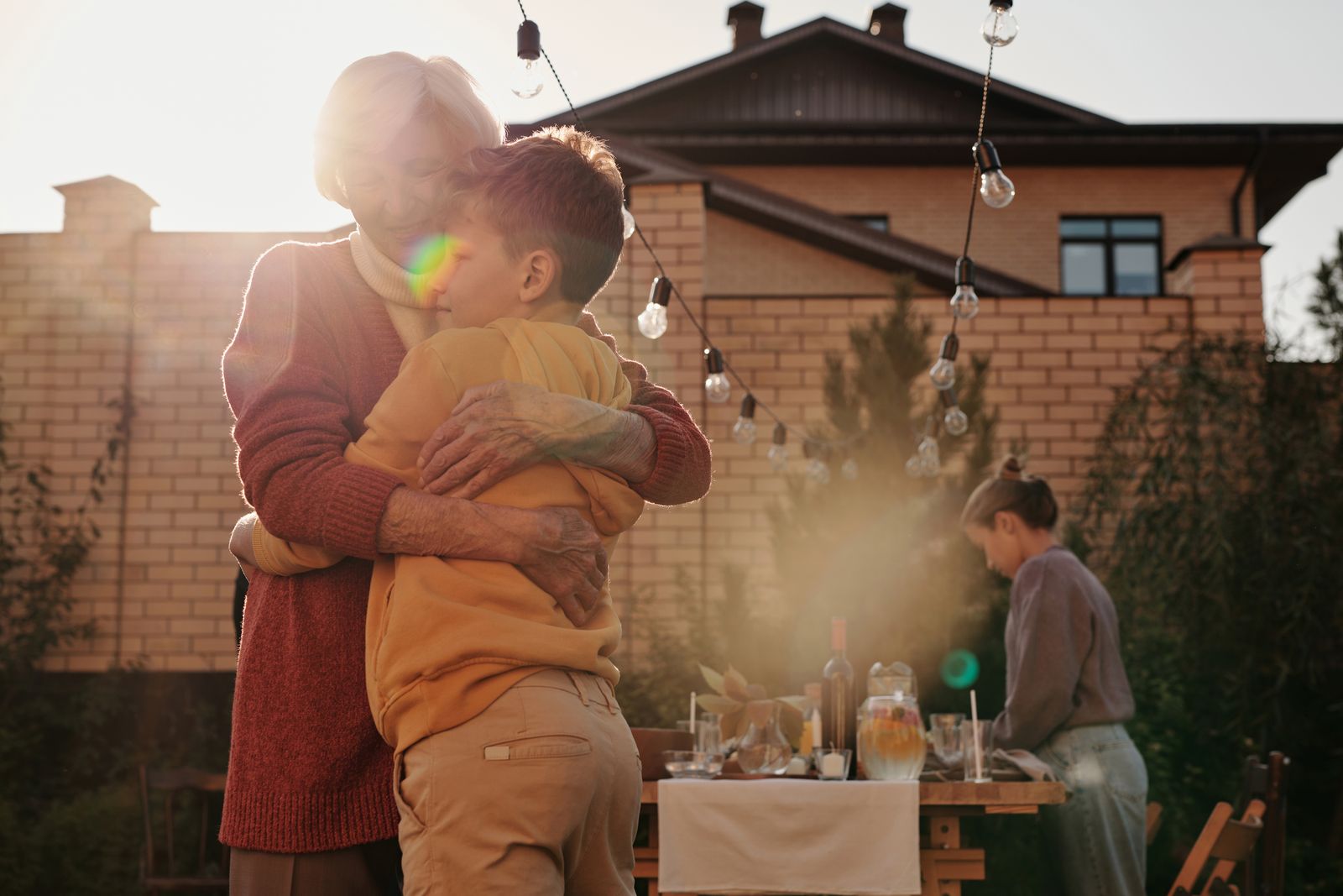 A grandmother warmly hugging a young boy at a backyard family dinner gathering, string lights overhead and a brick house behind, golden-hour sun flare; another family member sets the table in the…