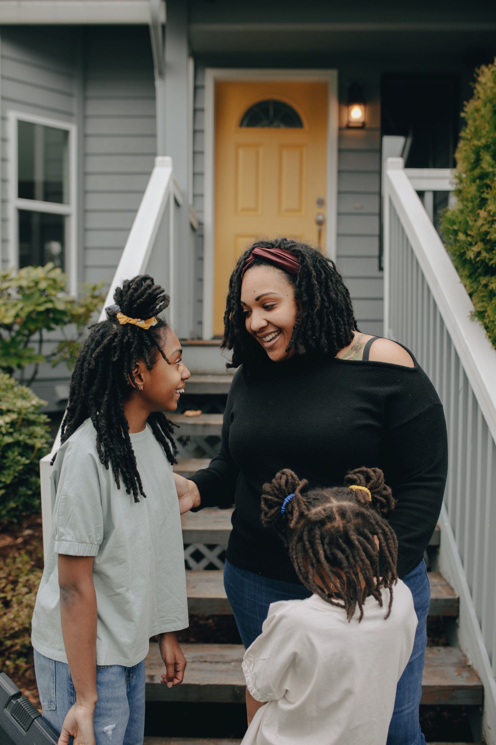A smiling mother laughing with her two daughters on the front porch steps of a gray suburban house with a bright yellow front door and porch light.