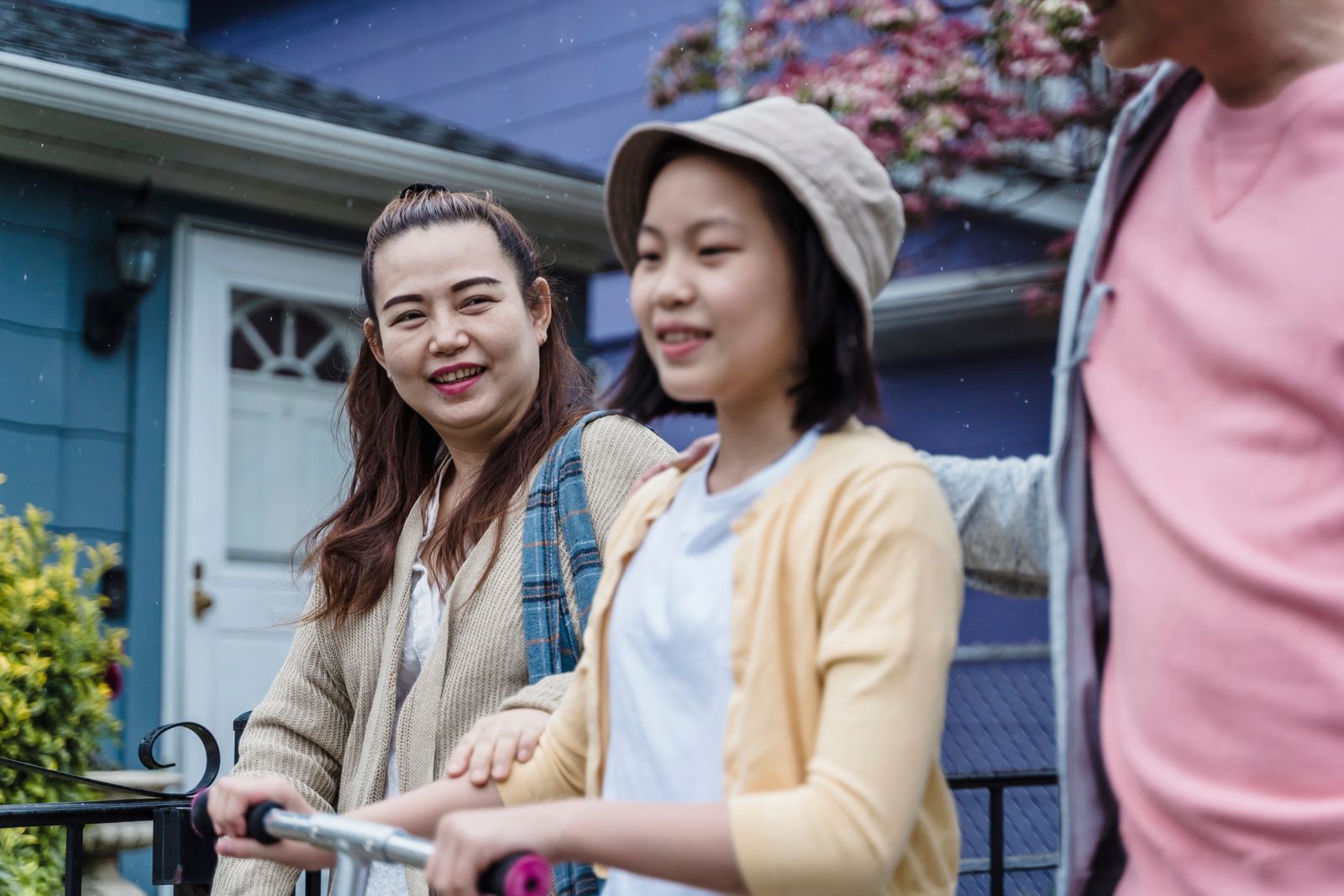 A smiling mother and her teenage daughter (plus father partly in frame) standing in front of a blue suburban house with a white front door and flowering tree; warm candid family moment.