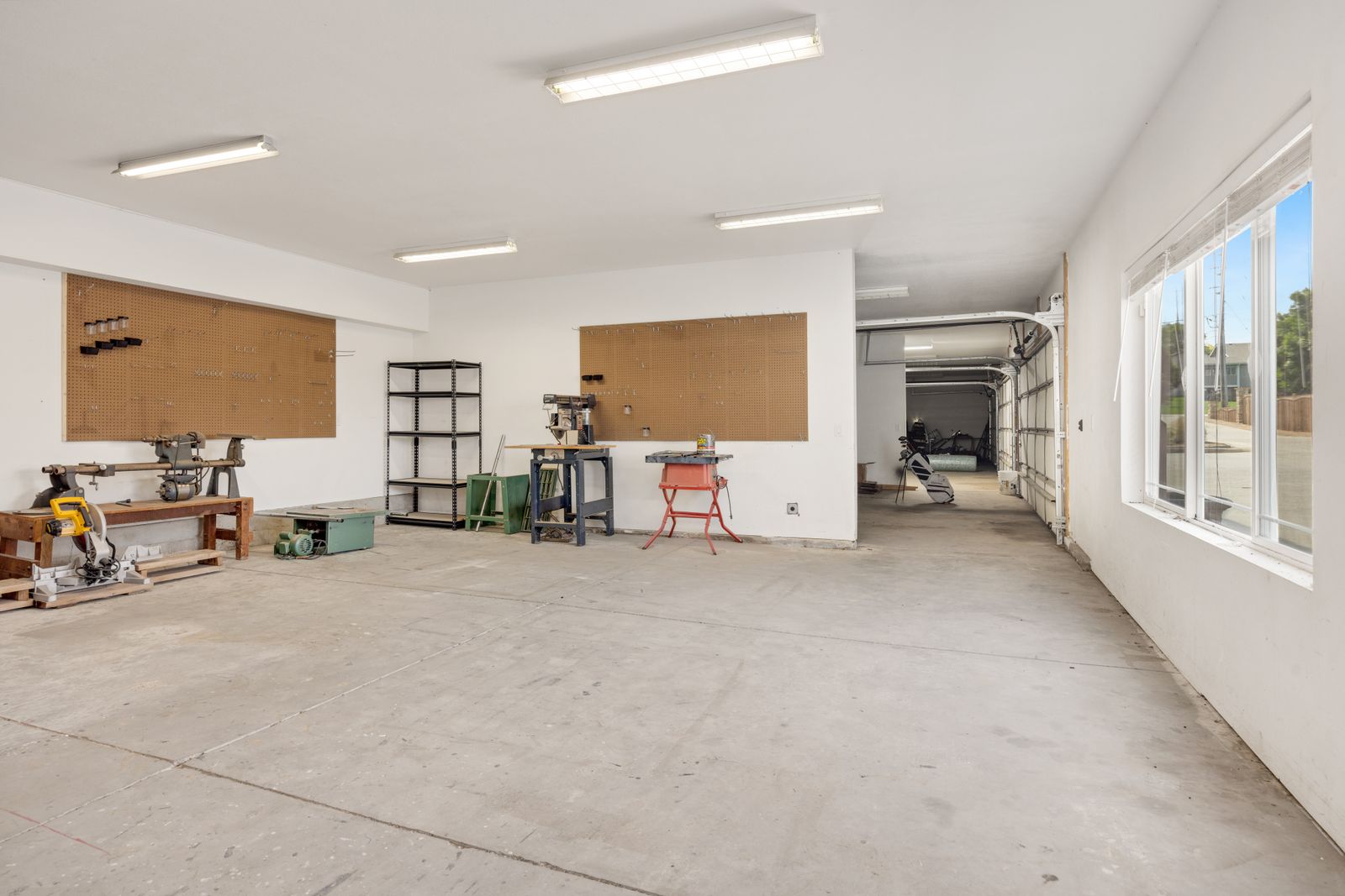 Bright, clean, spacious garage/workshop with white walls and ceiling, fluorescent lights, pegboards, a workbench and shelving on the left; at the far right a sectional garage door with its curved…