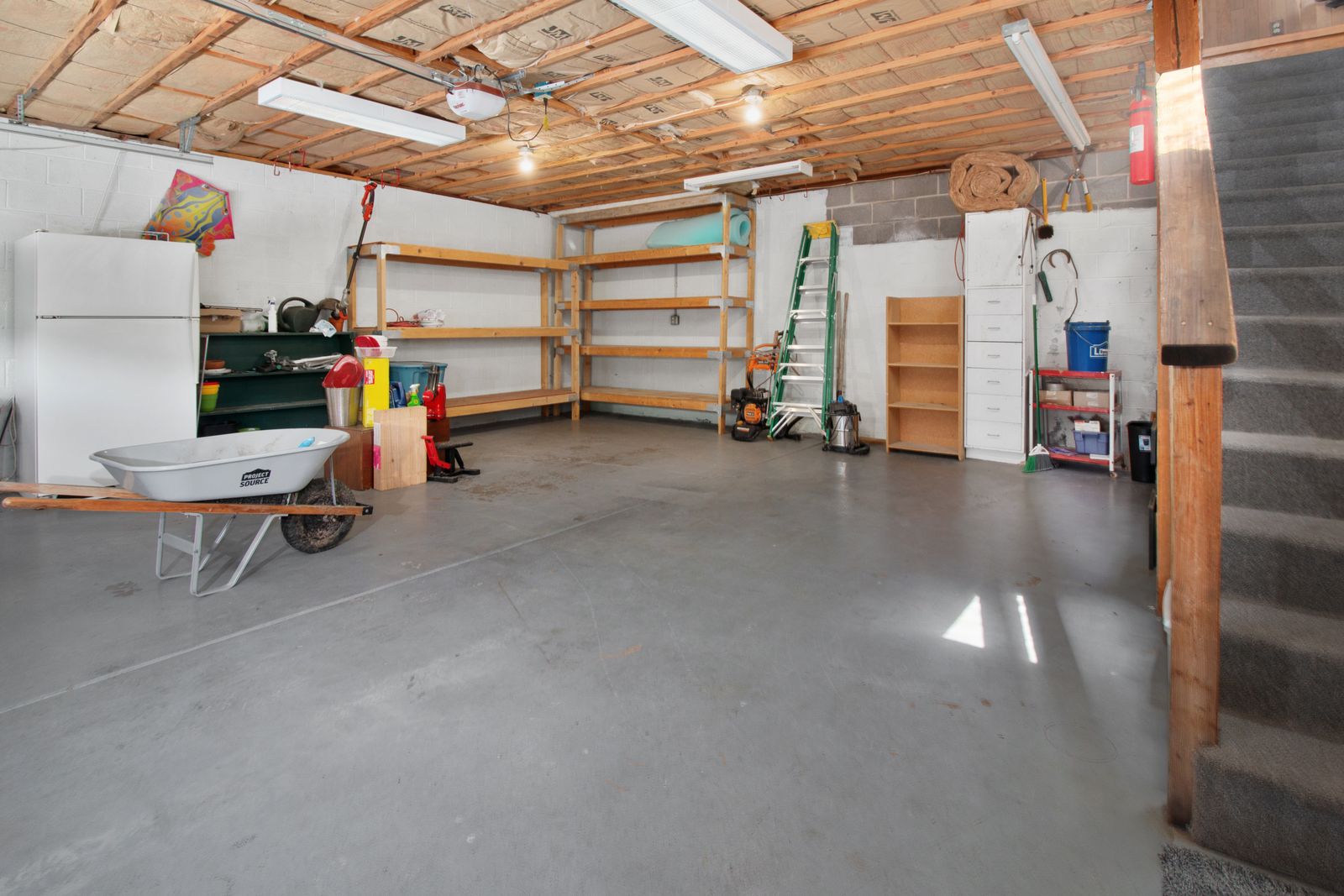 Interior of a residential garage with exposed wood ceiling joists; a chain-drive garage door opener is mounted to the ceiling rail running toward the camera, fluorescent shop lights, a white…
