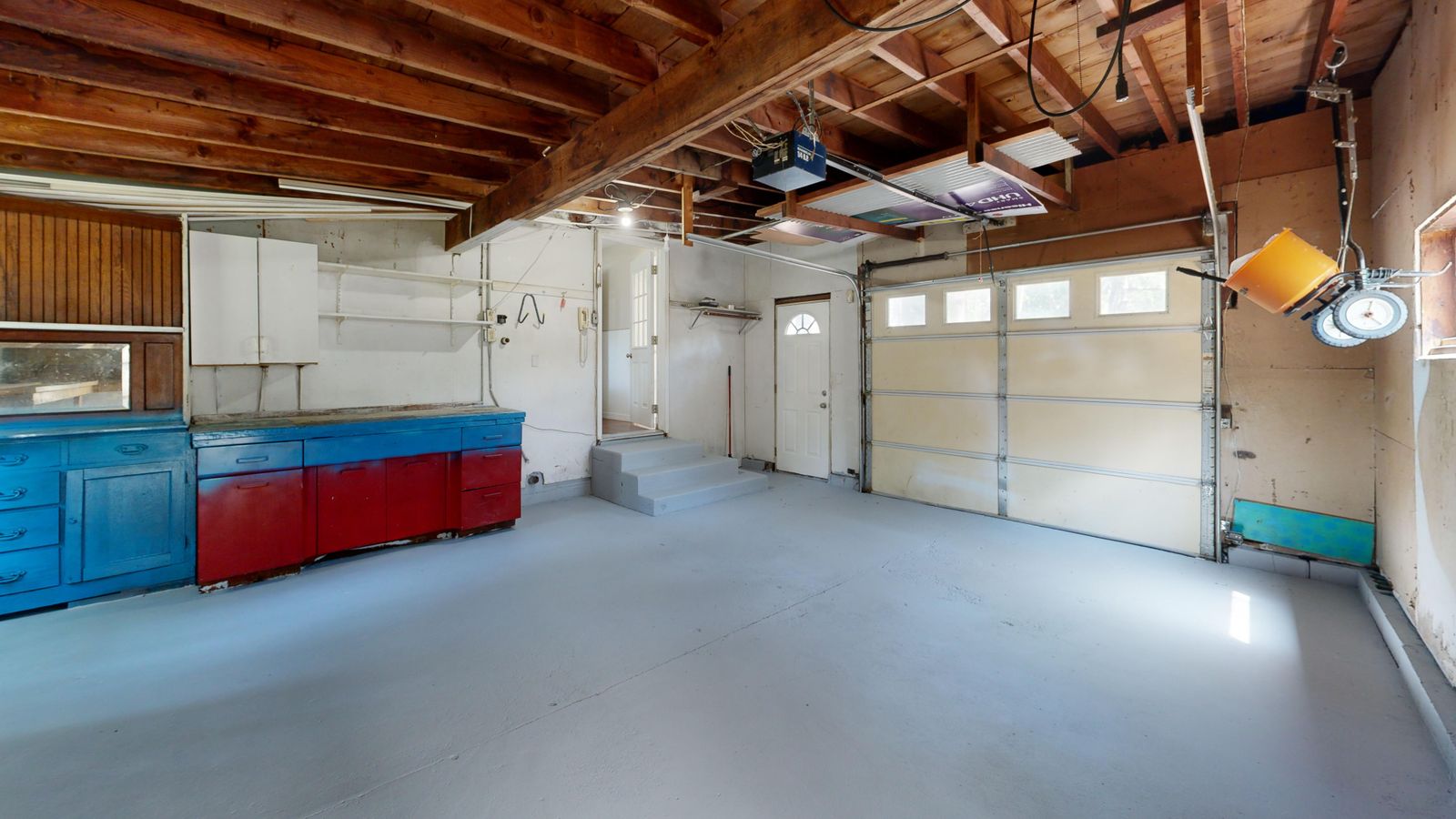 Inside a residential garage looking at a closed white sectional garage door with its horizontal overhead track and a ceiling-mounted opener visible on the exposed-joist ceiling; blue and red…