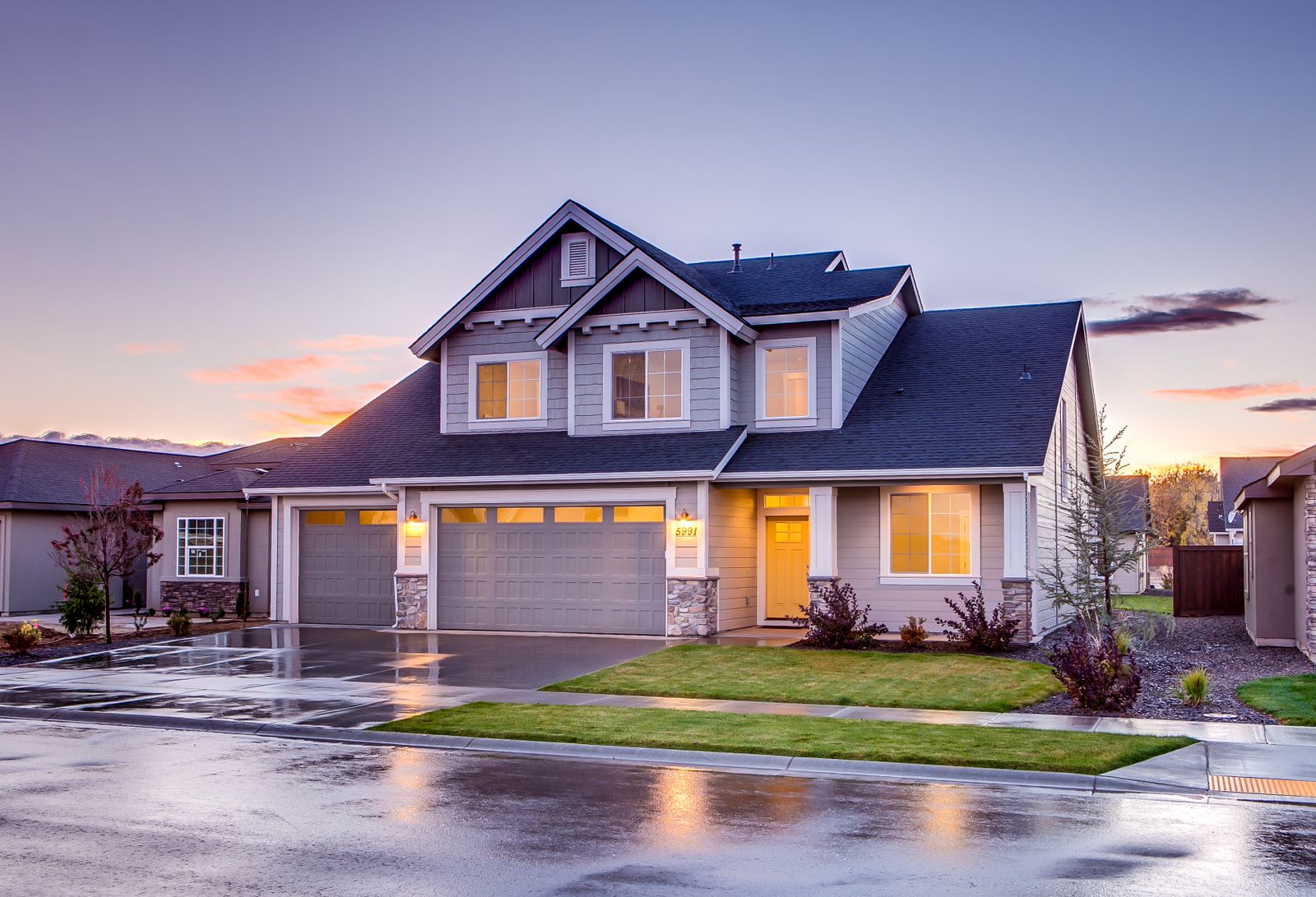 Gray craftsman two-story home with stone-base three-car garage, twilight sky with pink clouds, warmly glowing windows, wet driveway and street with reflections, neat lawn and landscaping.