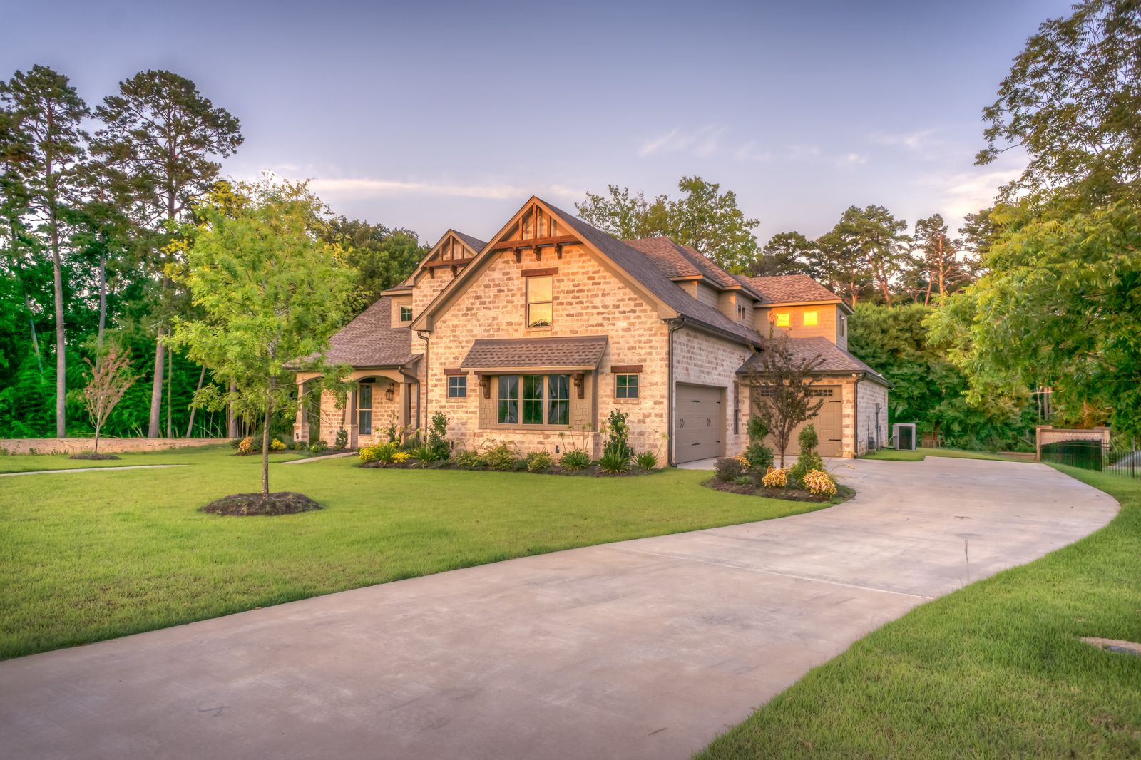 Light stone/brick two-story house with attached two-car garage, long curving concrete driveway sweeping across a green lawn, surrounded by trees at dusk with windows glowing warm.