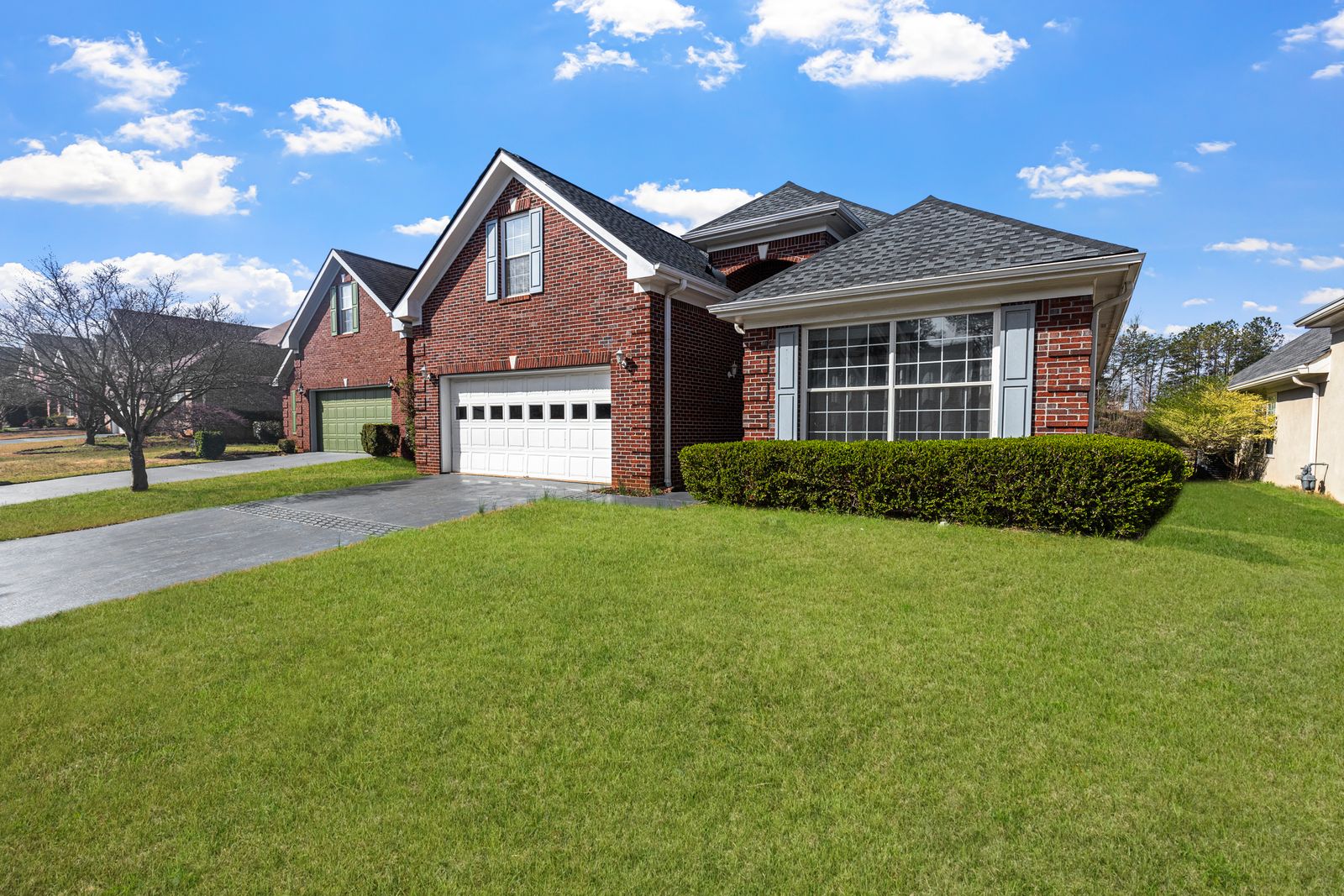 Charming red-brick suburban home with a white double garage door (plus a neighbor's green door at left), lush green lawn and bright partly-cloudy sky.