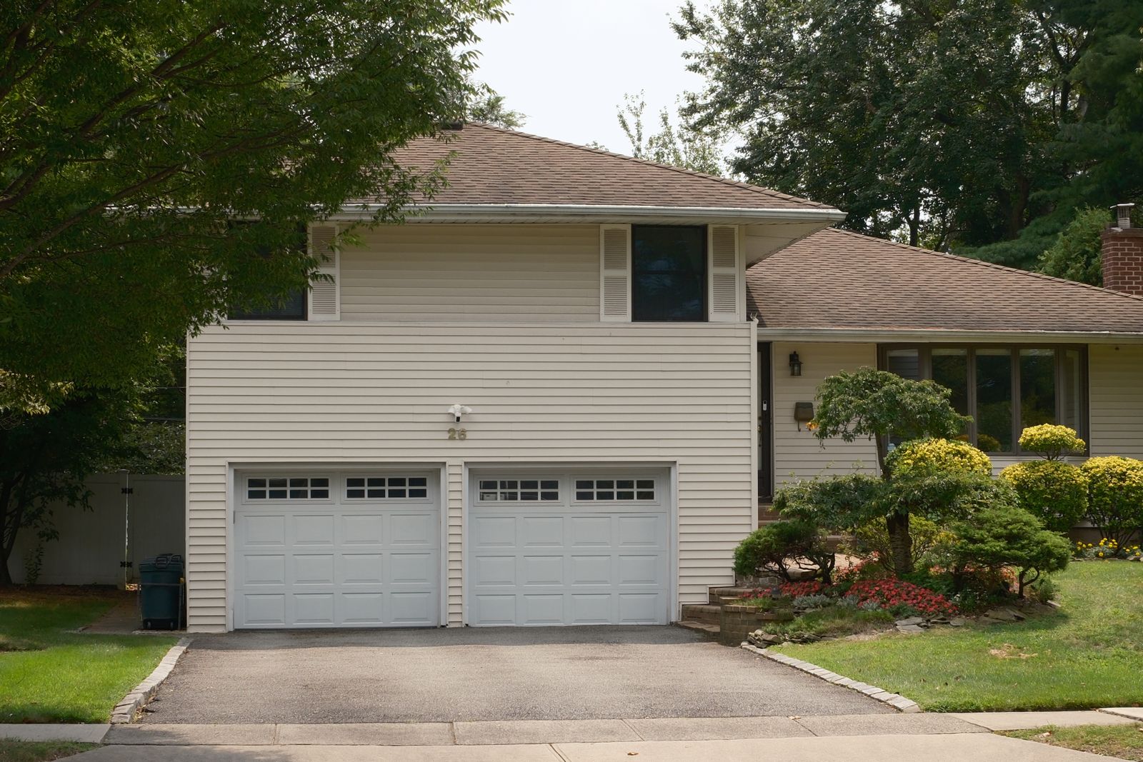 Tan split-level suburban home with two white sectional garage doors side by side, paved driveway, mature trees and landscaped front beds. Clear daylight.