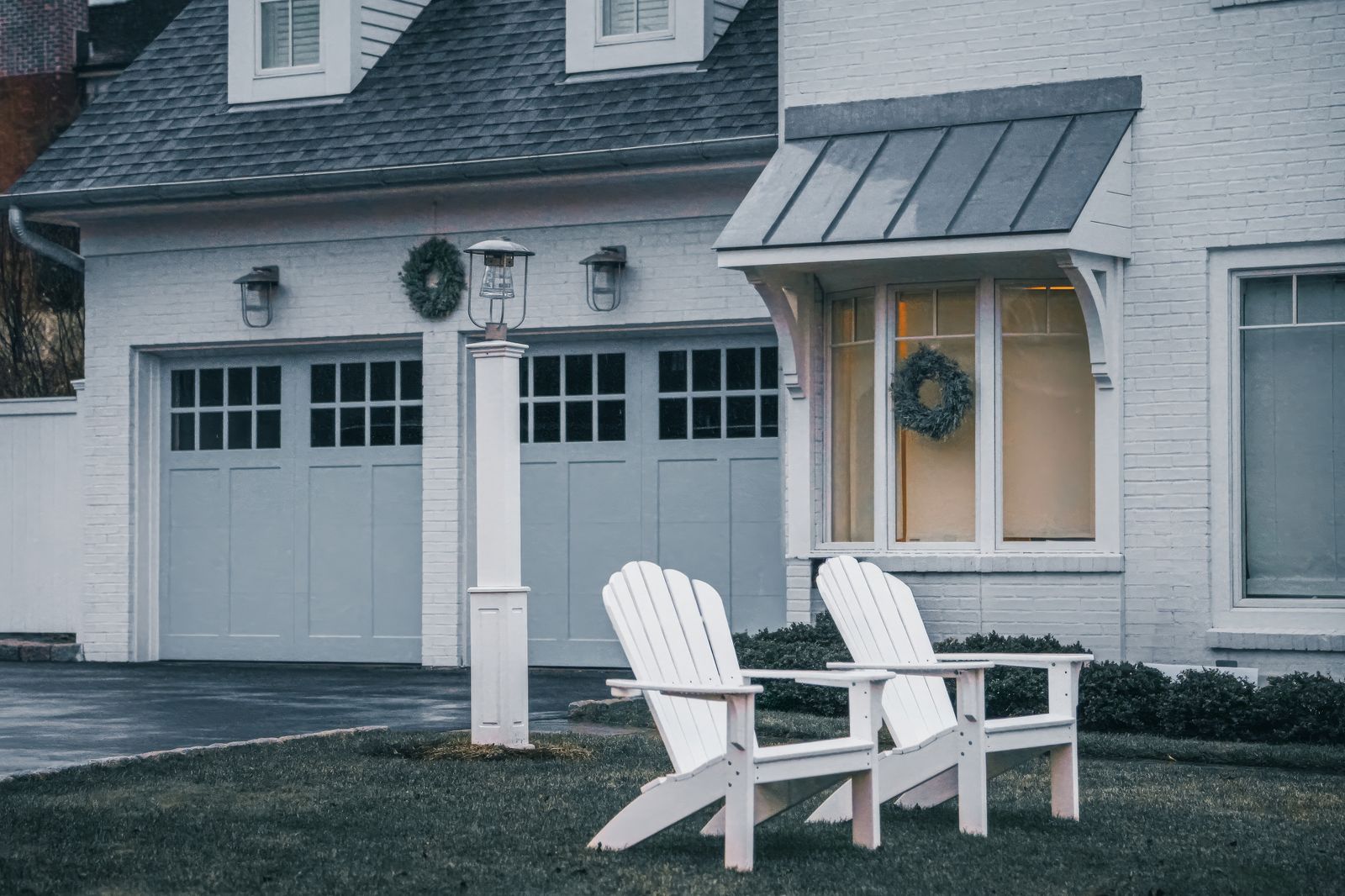White-painted home at dusk with two grey carriage-style garage doors topped by holiday wreaths and coach lamps, white Adirondack chairs on the lawn.