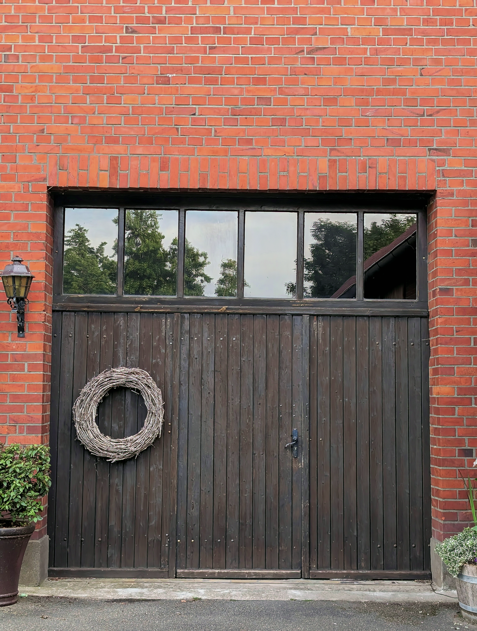 Close-up of a rustic dark-stained wood carriage-style garage door with a window row, set in a red-brick wall, decorative wreath and coach lamp beside it.
