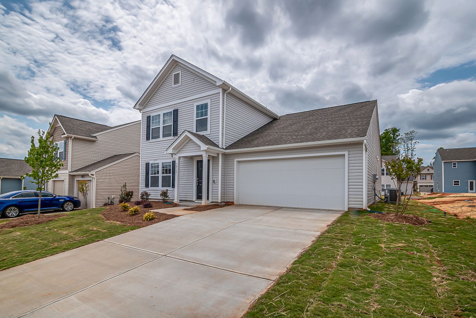 Light-gray two-story suburban home with a white double garage door, wide concrete driveway and a blue sedan parked at left, dramatic cloudy sky.