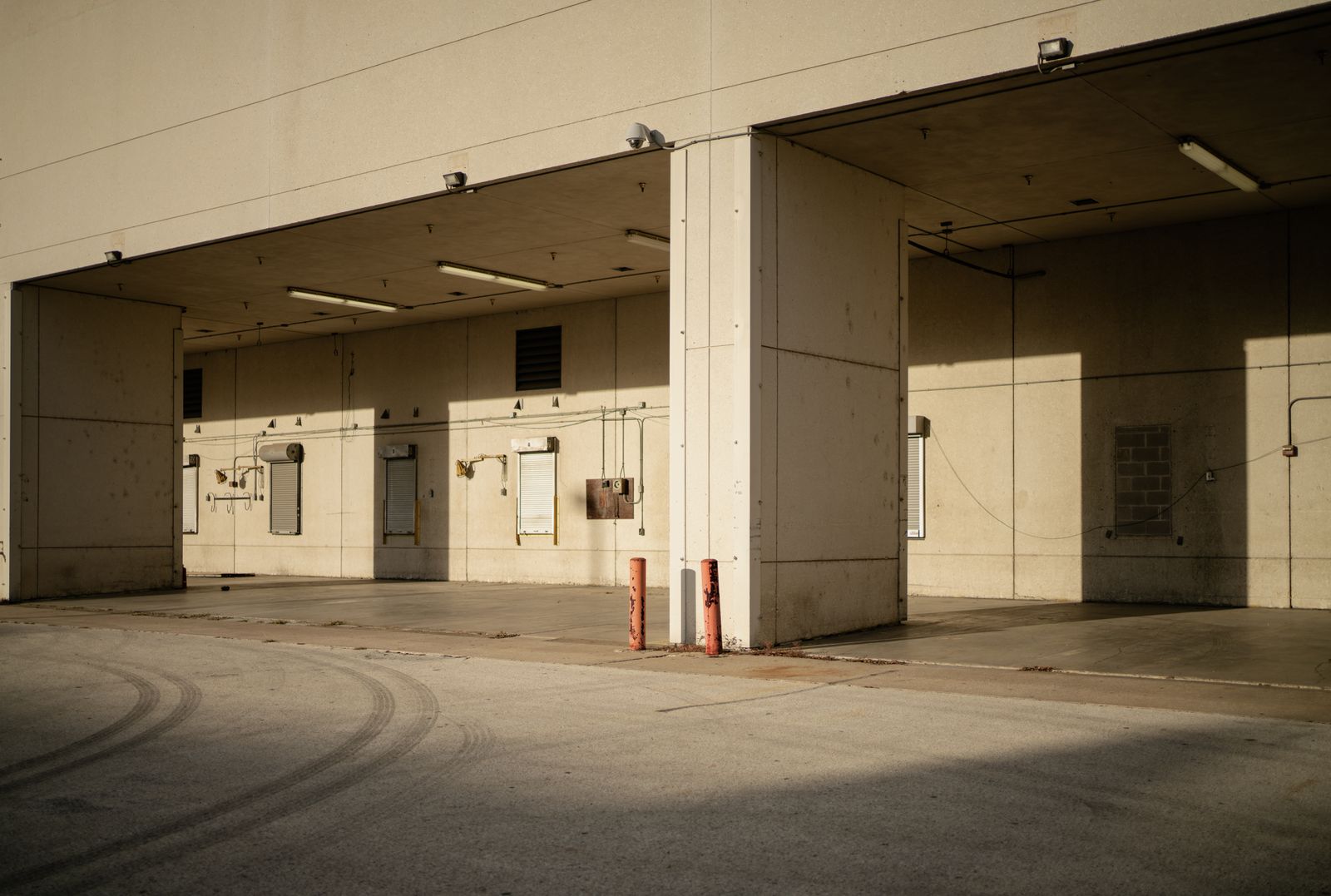 Drive-under covered loading dock with several recessed bays and roll-up doors set into a tan concrete building, warm golden side-light, empty driveway with tire marks in front.