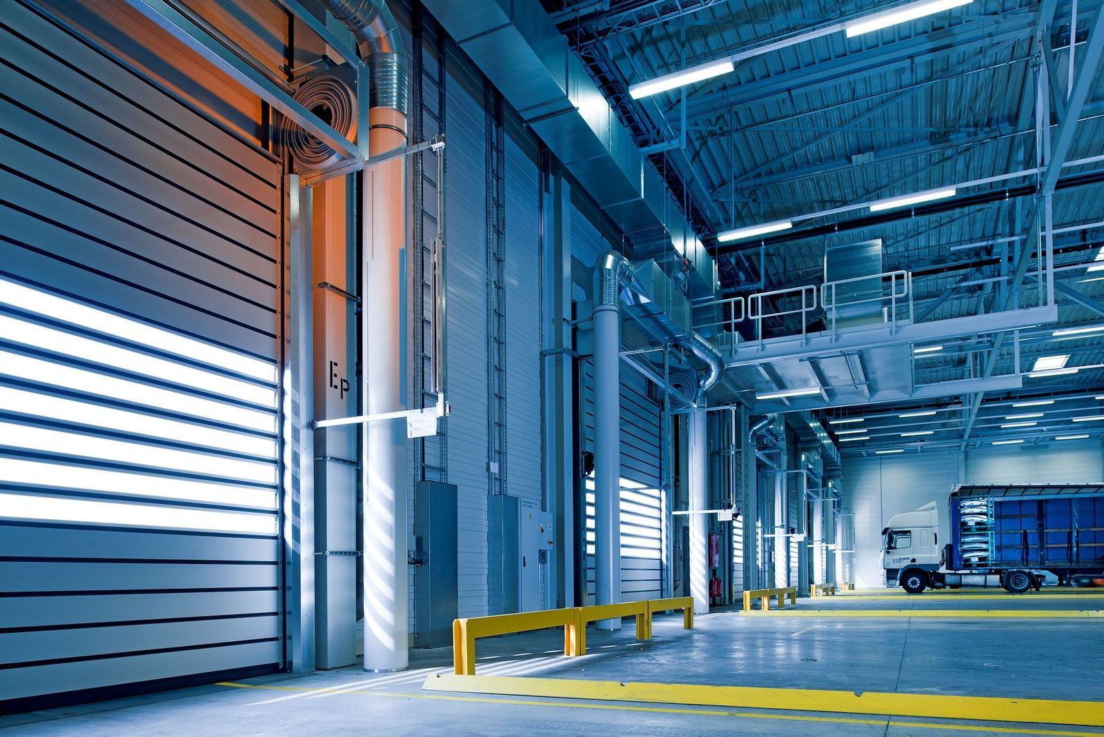 Interior of a modern industrial warehouse: a row of sectional/roll-up dock doors along the left wall (one open with daylight), overhead lighting and ductwork, a white truck cab with trailer parked…