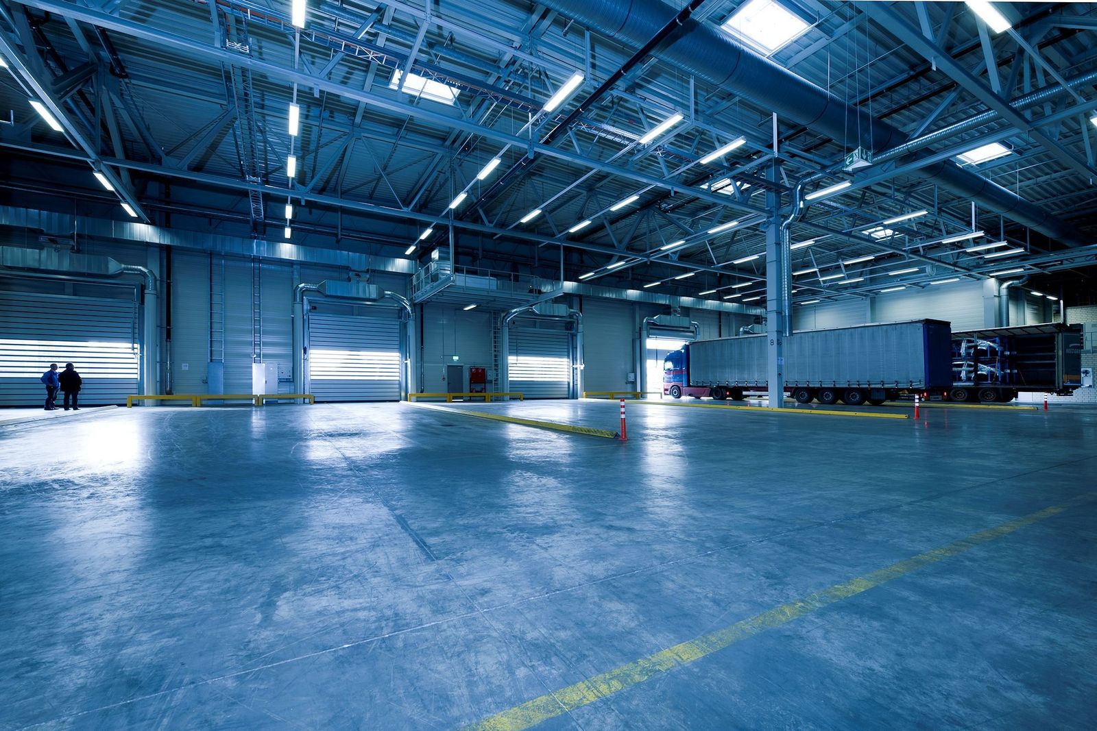 Wide interior of a large blue-lit warehouse with a line of roll-up dock doors on the left, a blue tractor with a curtain-side trailer parked at the bays, polished concrete floor with yellow lane…