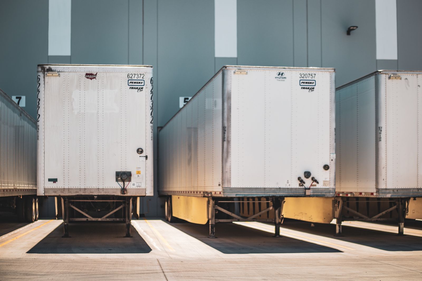 Two white semi box trailers backed squarely into numbered dock bays against a gray warehouse wall, dock seals visible, bright daylight.