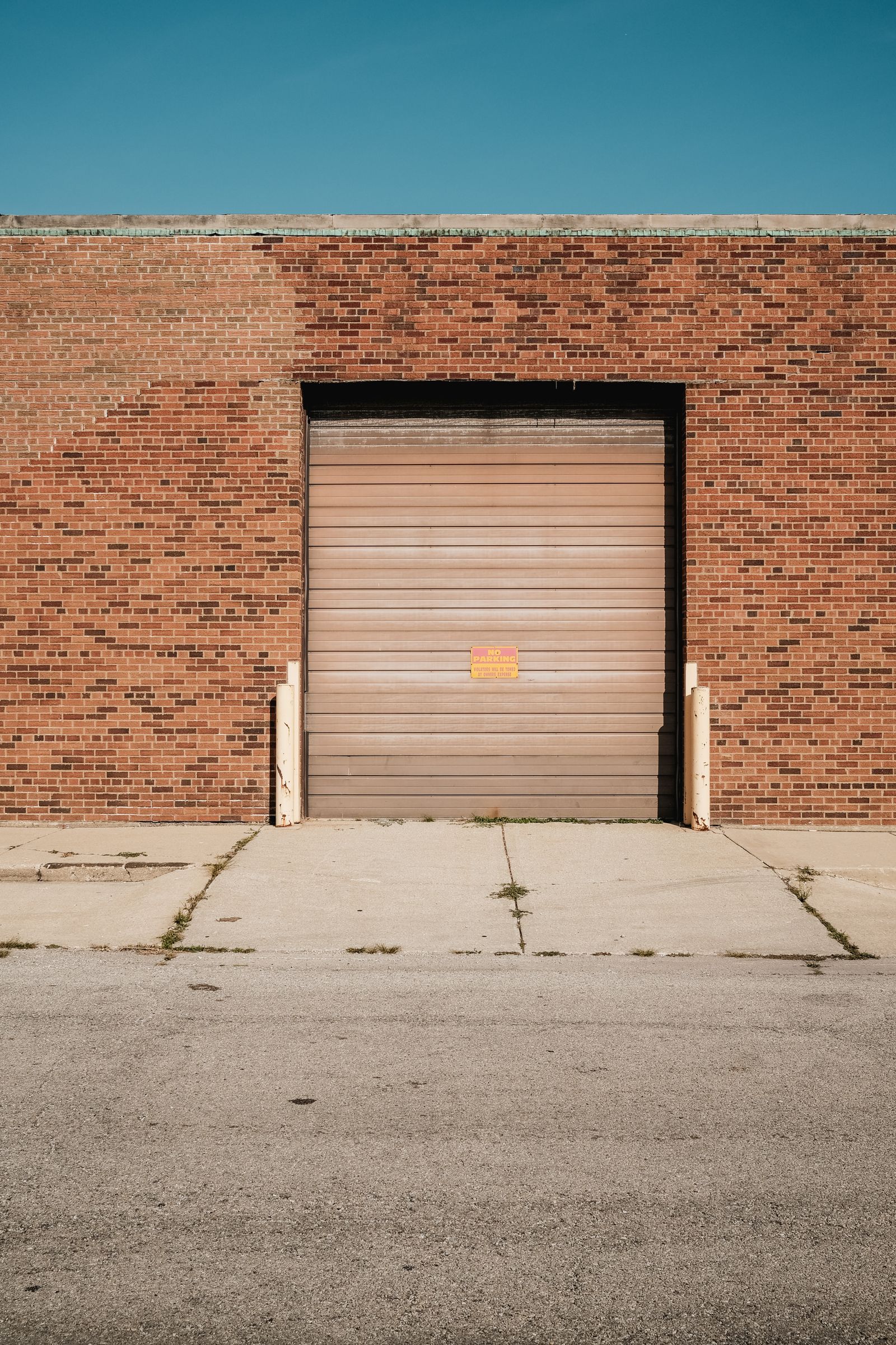 Single beige commercial roll-up garage door centered in a red-brick warehouse wall under clear blue sky, concrete pad and street in foreground.