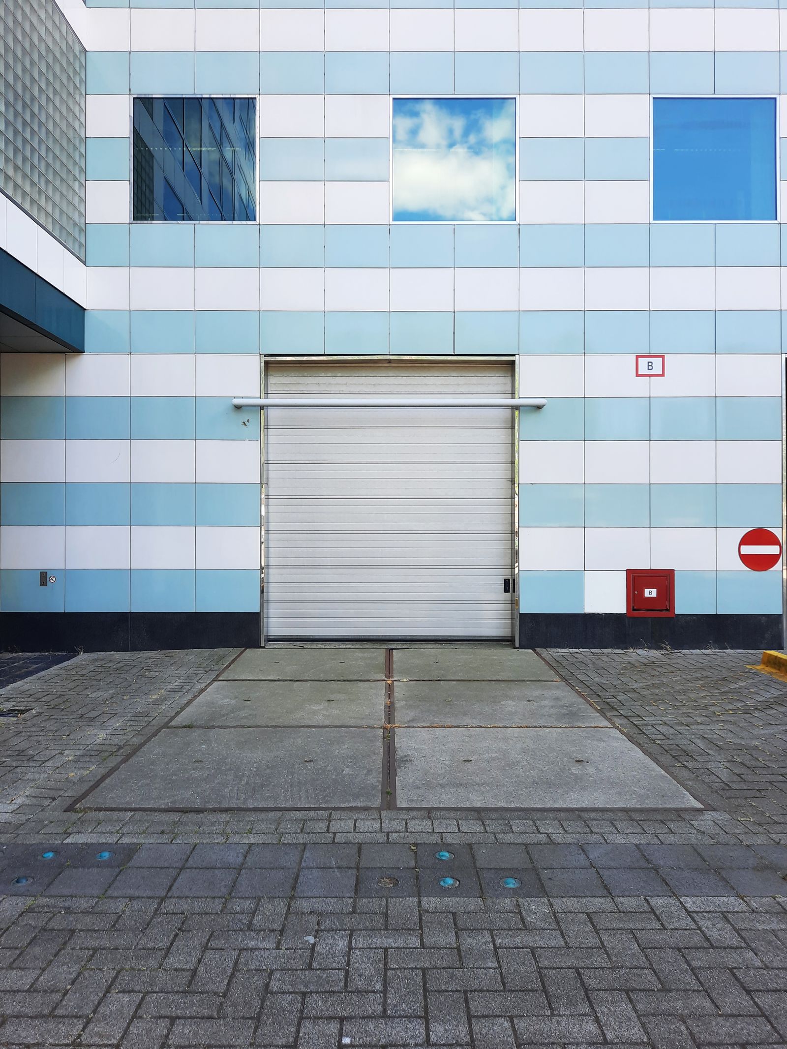 White metal roll-up overhead door set into a modern blue-and-white tiled warehouse facade with a one-way sign and fire box to the right, paved apron in foreground.