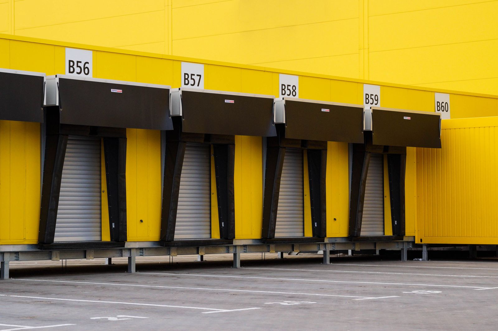 Row of bright-yellow industrial loading-dock bays numbered B56-B60, each with a black dock shelter/canopy and a metal roll-up door behind it, empty concrete apron in front.