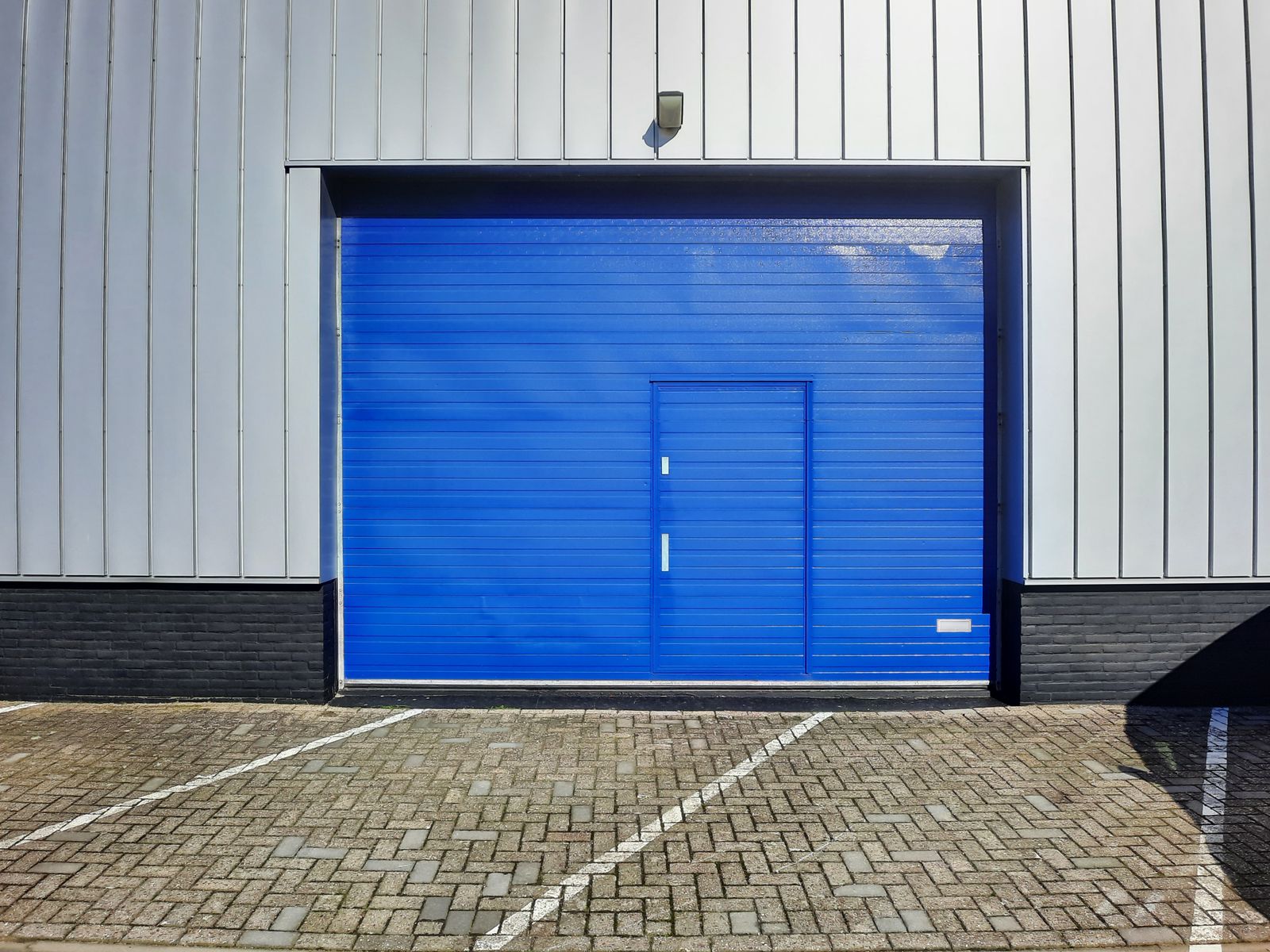 Clean modern commercial sectional roll-up door painted bright blue with an integrated blue pass-door, set in a gray metal-clad warehouse wall, paver driveway in foreground, bright sun.