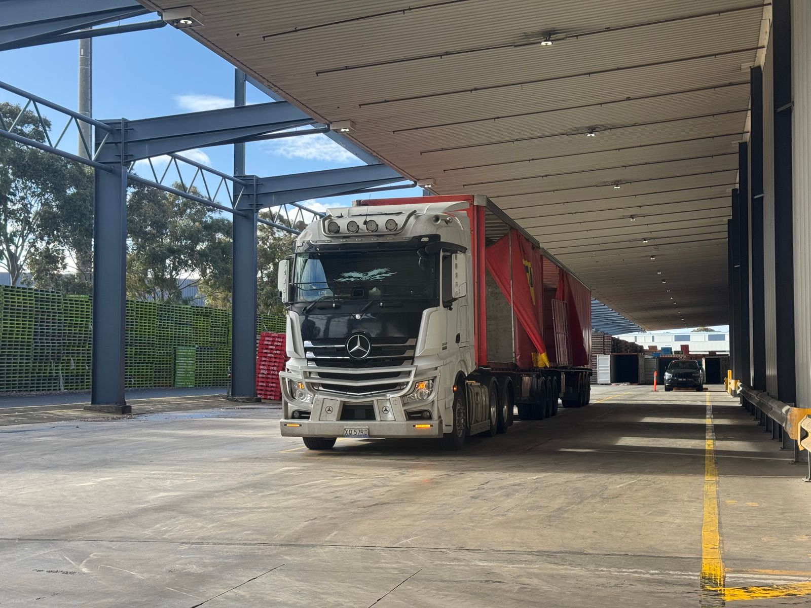 White Mercedes-Benz Actros semi tractor with a red curtain-side trailer parked under a steel-framed covered loading-bay canopy, blue sky and greenery at left.