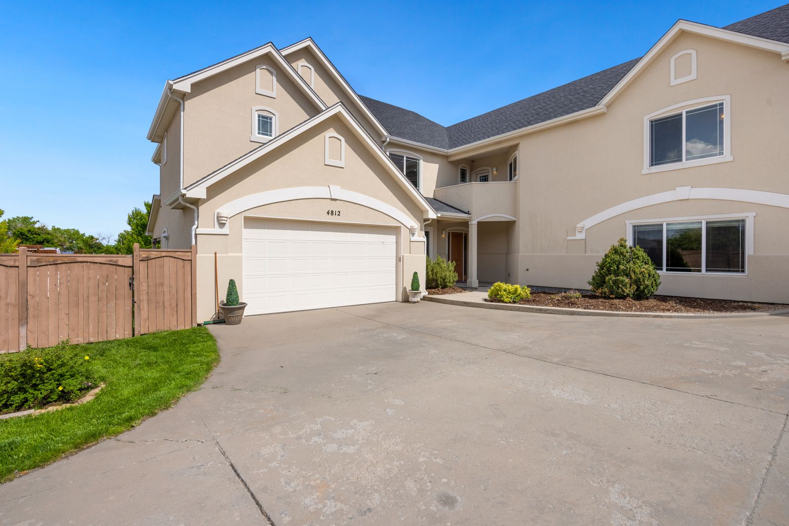 Two-story tan stucco home with arched entry and a white double-wide sectional garage door at the end of a curving concrete driveway; cedar privacy fence at left, trimmed shrubs, deep blue sky.