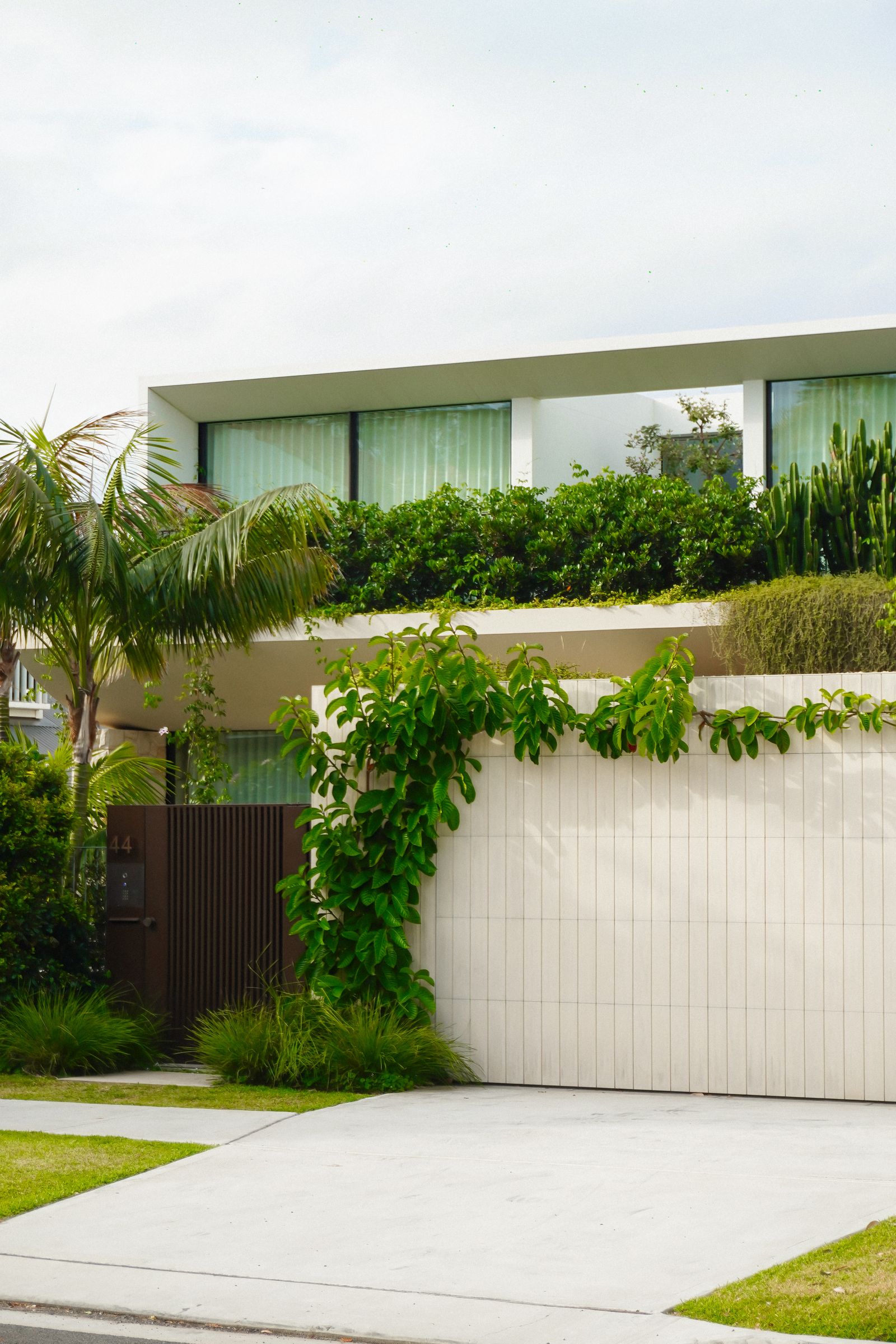 Two-story flat-roofed modern white home with a planter ledge of greenery and climbing vines spilling over a clean white sectional garage door; concrete driveway in foreground, overcast bright sky.