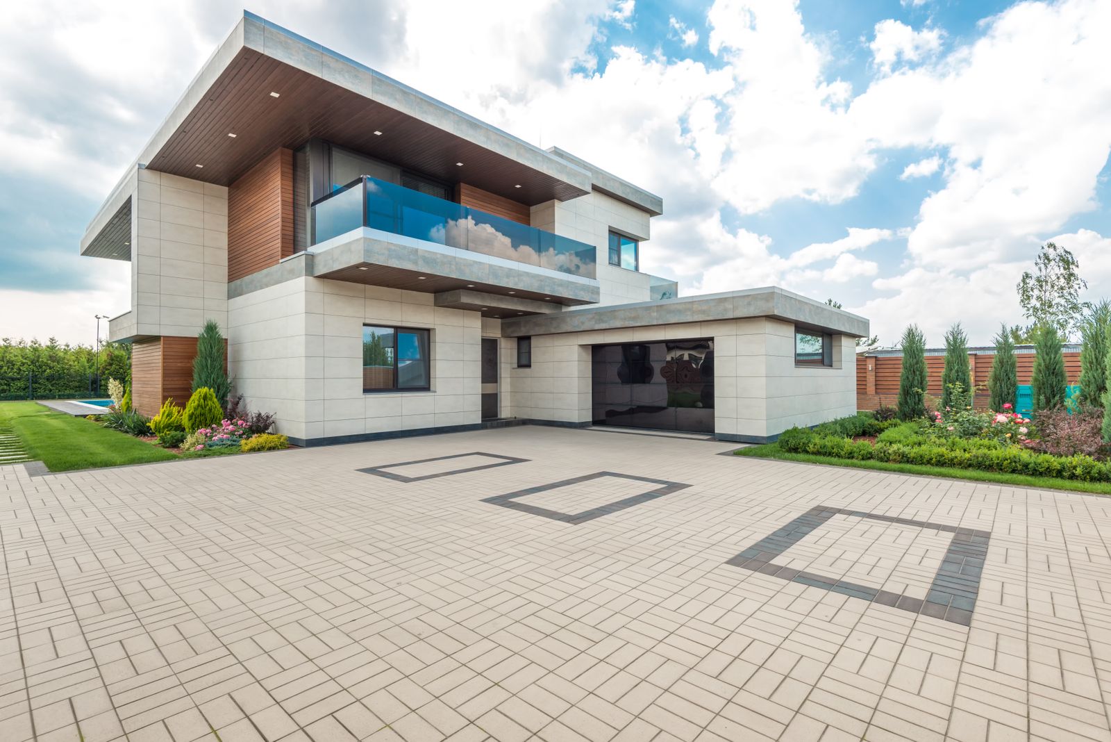 Wide exterior of a luxury contemporary home with cantilevered flat roof, cream stone-tile facade, warm wood accents and glass balcony; a large dark tinted-glass garage door is integrated into the…