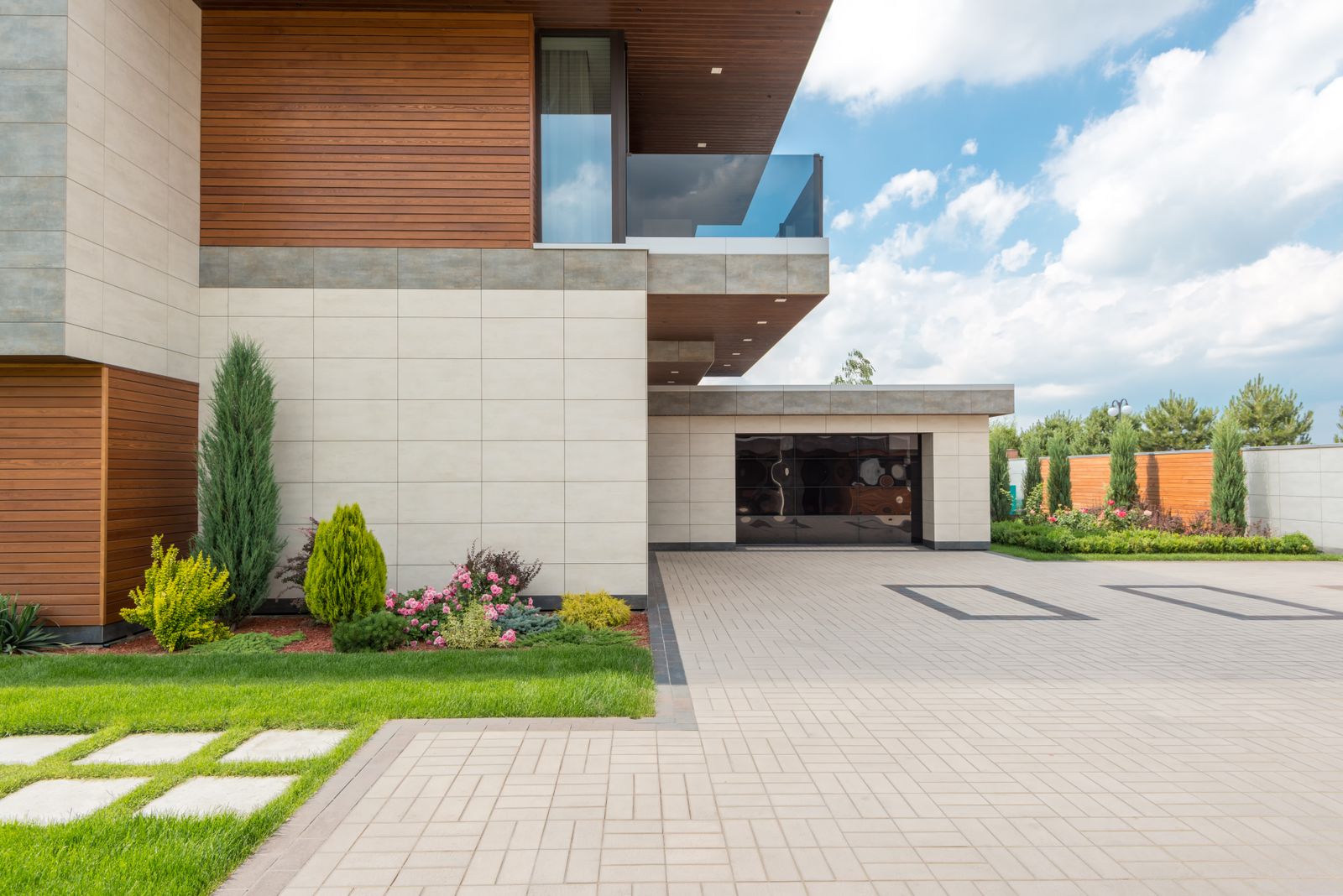 Close exterior detail of the same luxury modern home: cream stone-tile and horizontal-wood facade, second-floor glass balcony, and a recessed dark glossy garage door, with a paver driveway,…