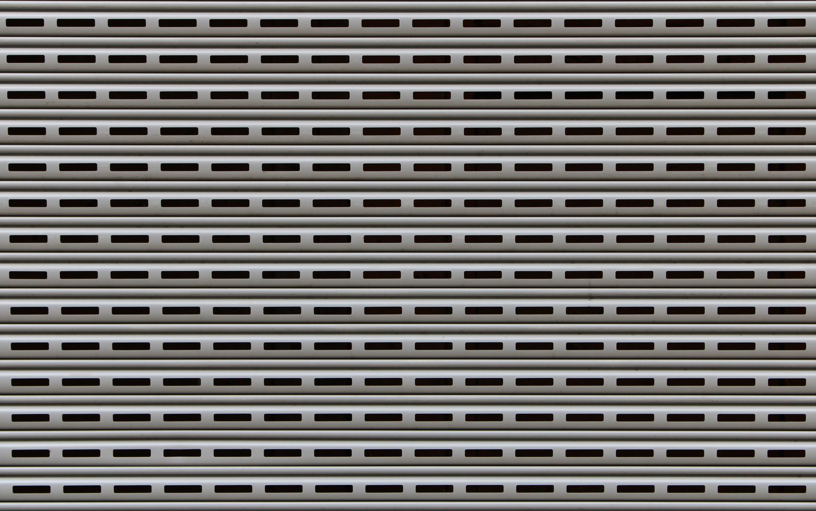 Full-frame close-up of a perforated/vented grey rolling security shutter, horizontal slats with rectangular vent slots.