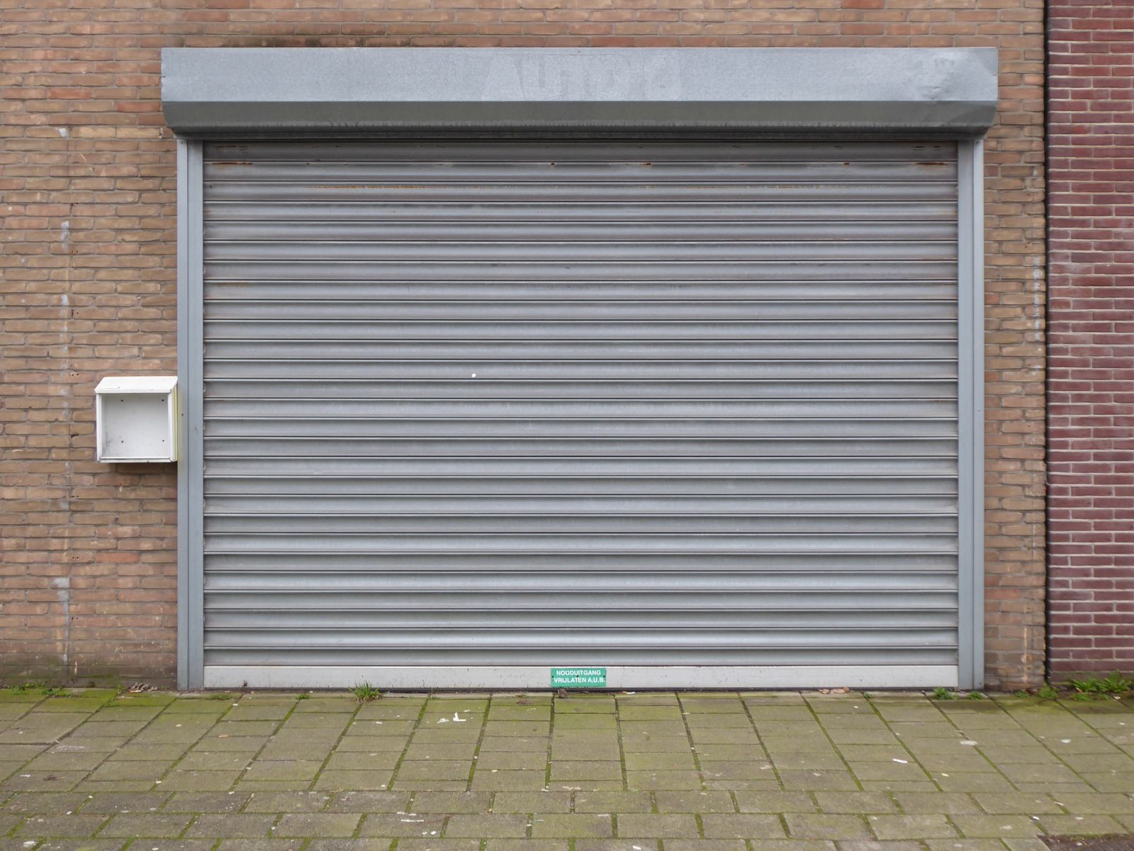 A grey commercial rolling steel shutter door (closed) with a hood box on top, set into a red-brick wall above paver sidewalk.