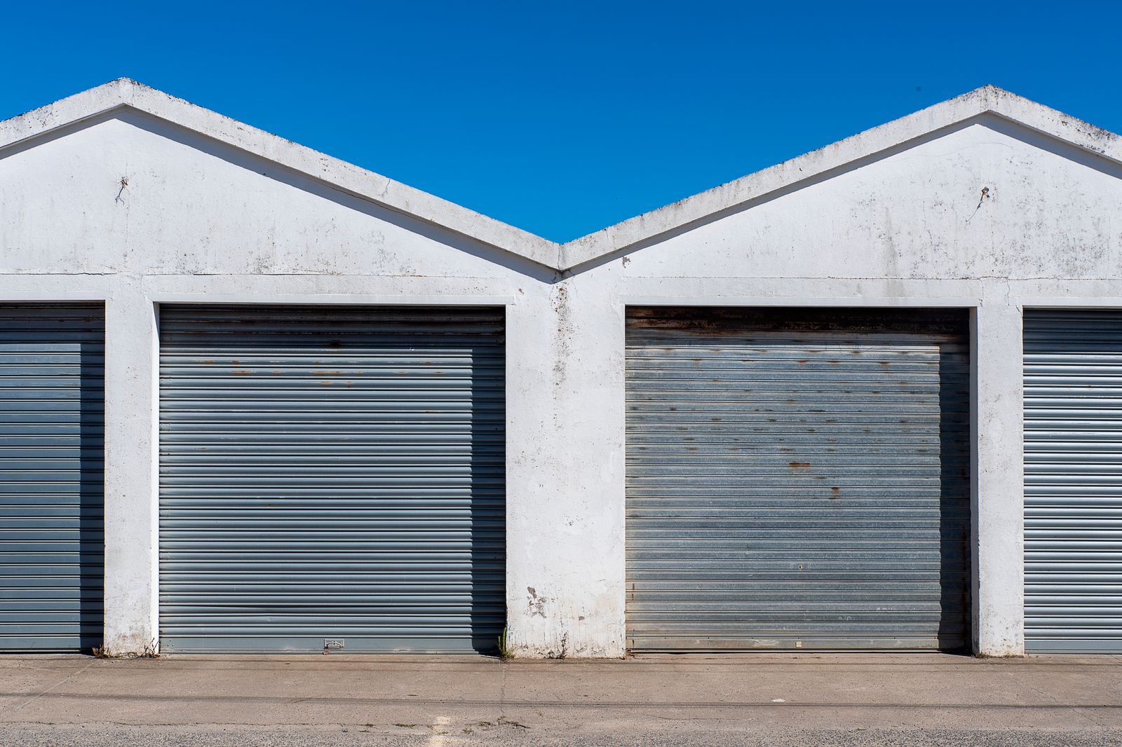 Two side-by-side commercial roll-up storage units with light-grey corrugated rolling steel doors set in white gabled concrete bays, under a clear deep-blue sky, asphalt apron in front.