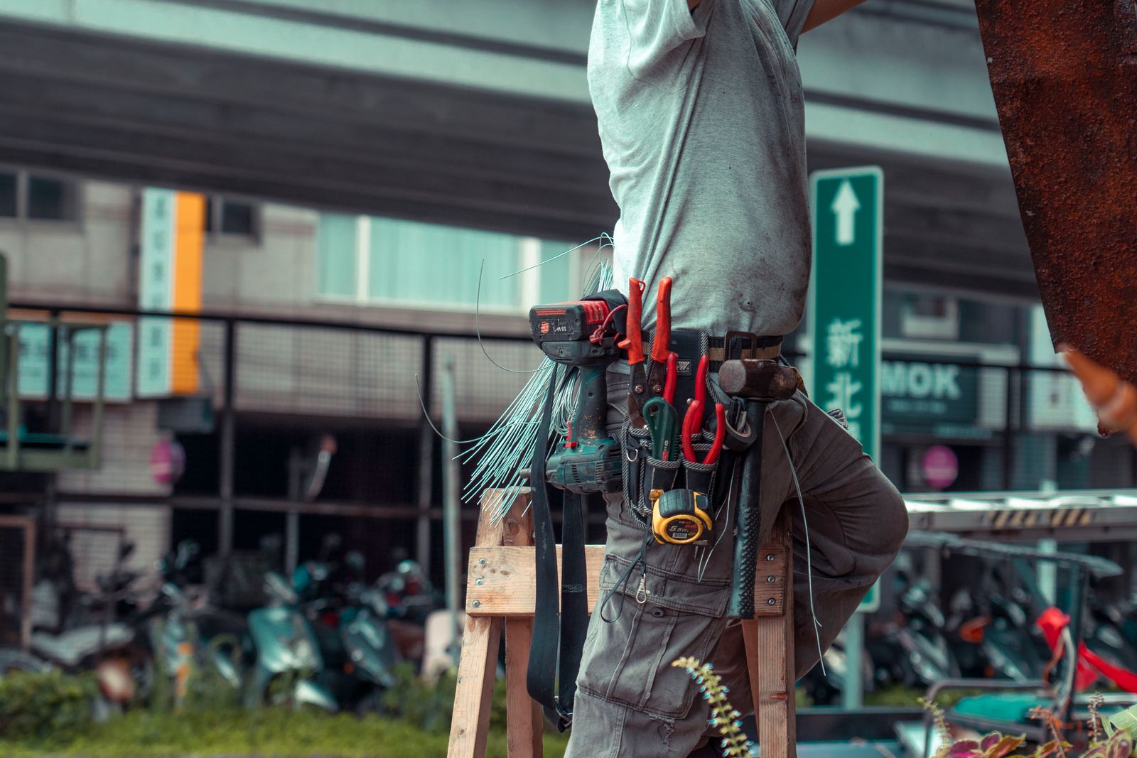 Close-up of a tradesman's torso and hip showing a fully loaded tool belt (drill, pliers, screwdrivers, tape measure) while he straddles a wooden sawhorse/ladder in an urban setting.