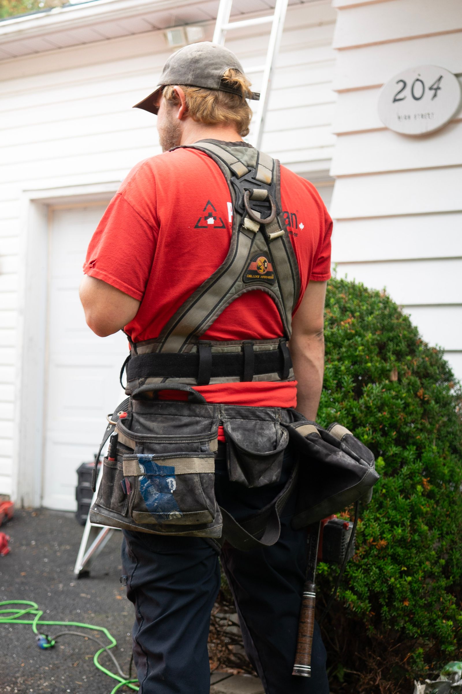 Rear/side view of a male technician in a red tee wearing a heavy-duty tool belt and a safety harness loaded with pouches and tools, standing in front of a white residential garage door (house…
