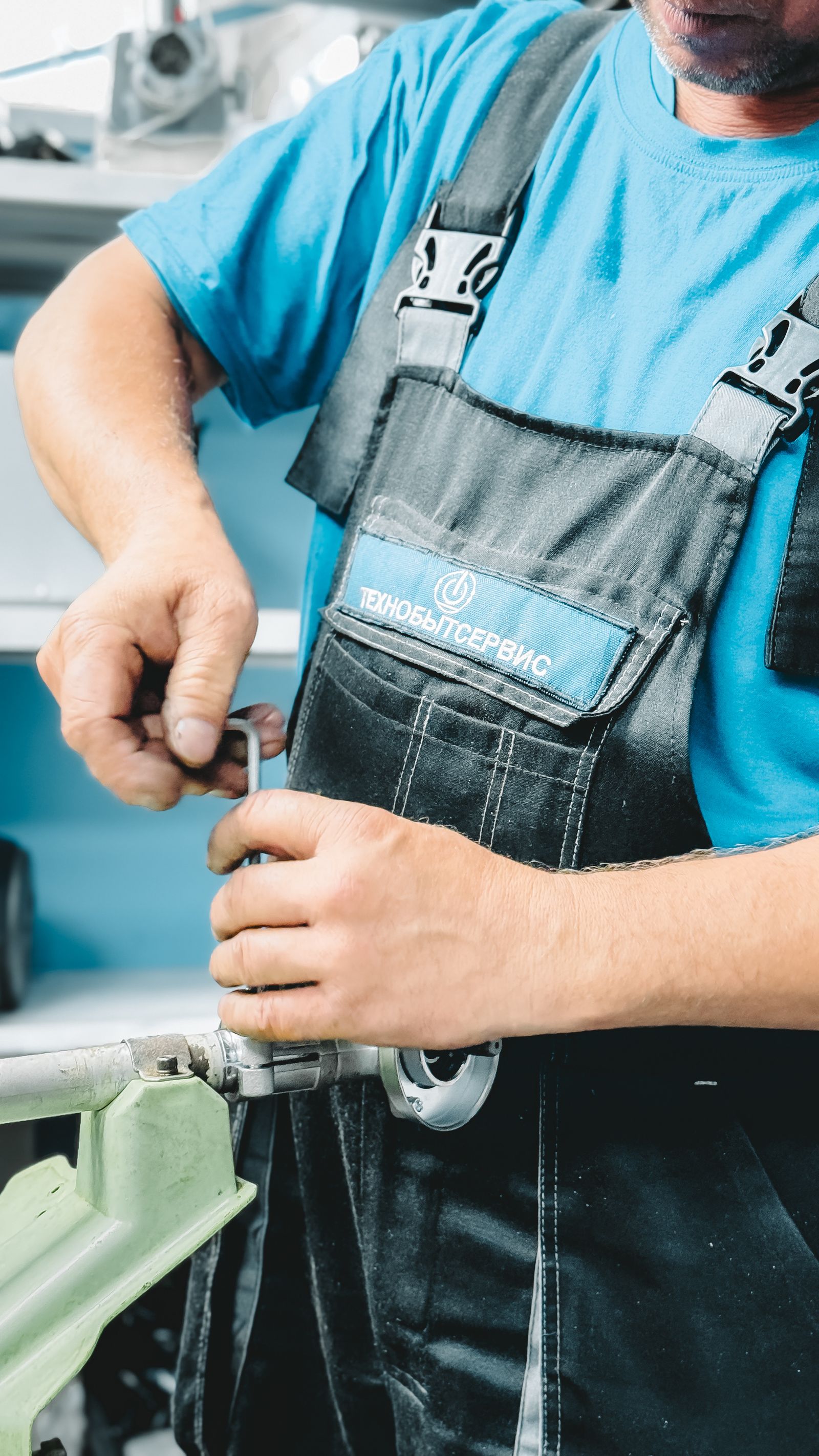 Torso-framed shot of a technician in blue shirt and work overalls using both hands to fix a part with a small tool at a workbench; hands and grip are the focal point.