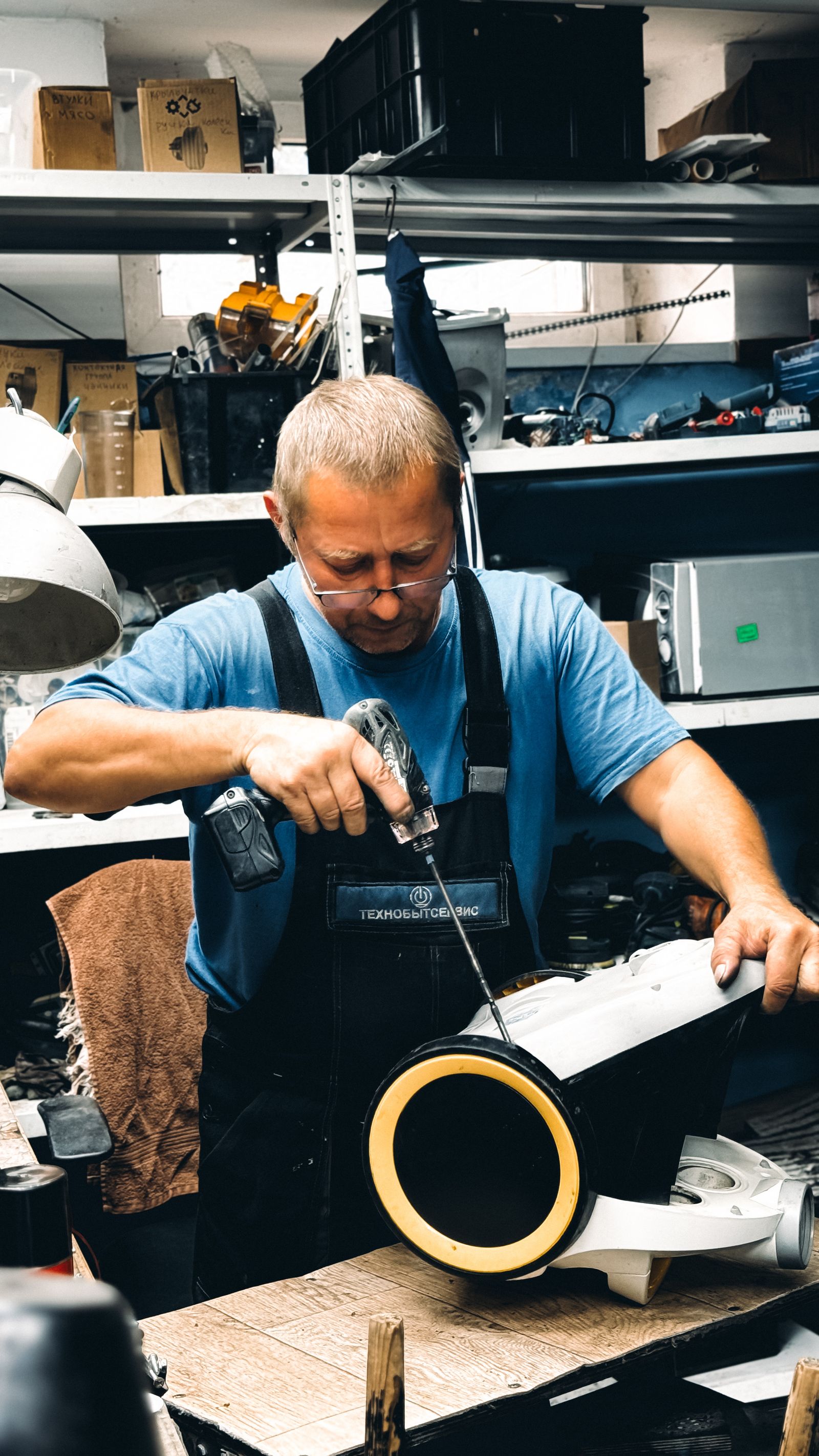 Technician in glasses, blue shirt and work overalls actively driving a cordless drill/driver into a part he holds with his other hand, focused on the task, in a repair workshop.