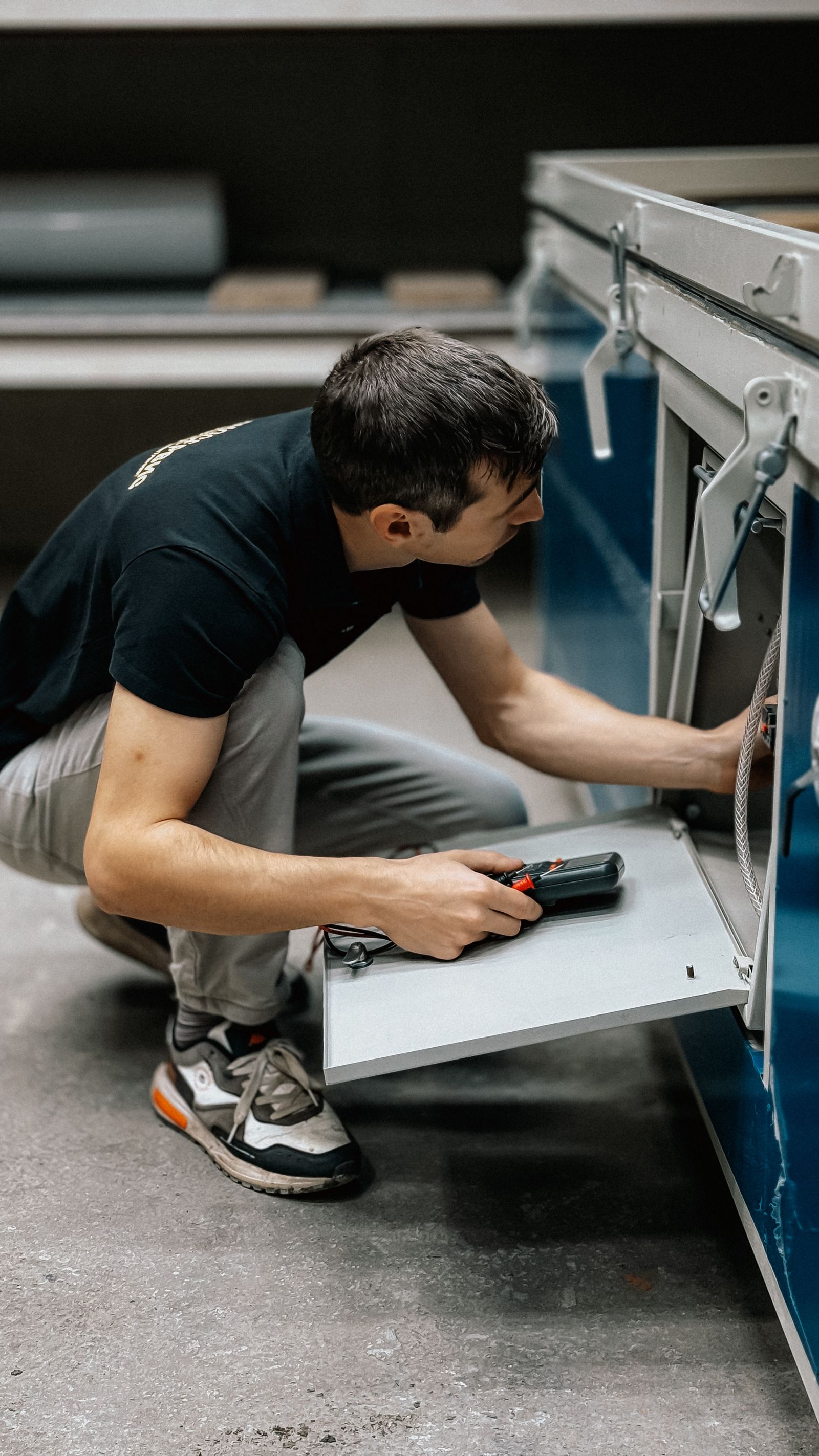 Technician in a black tee and gray pants crouched low, using a small hand tool to work on a cabinet/cable mechanism with a metal cable visible; clean modern interior.
