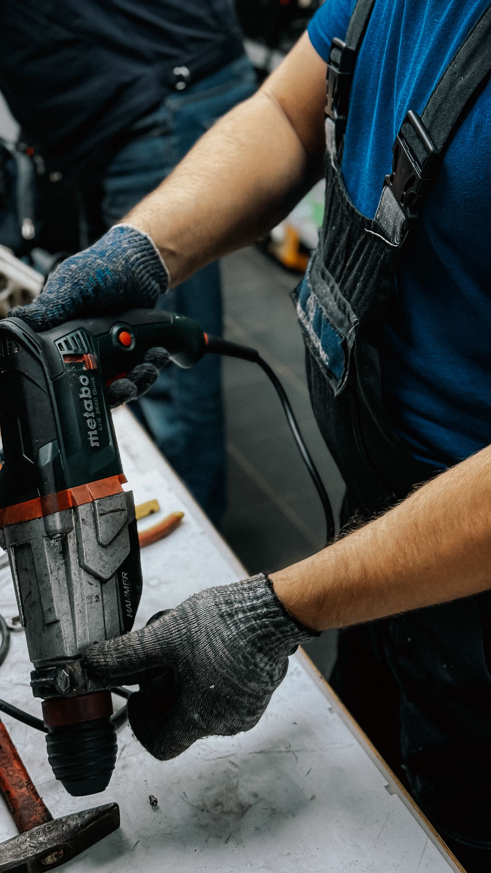 Tight close-up of a technician's hands in gray work gloves gripping a Metabo rotary hammer drill over a workbench, second gloved hand steadying the bit; blue shirt and work overalls visible at top.