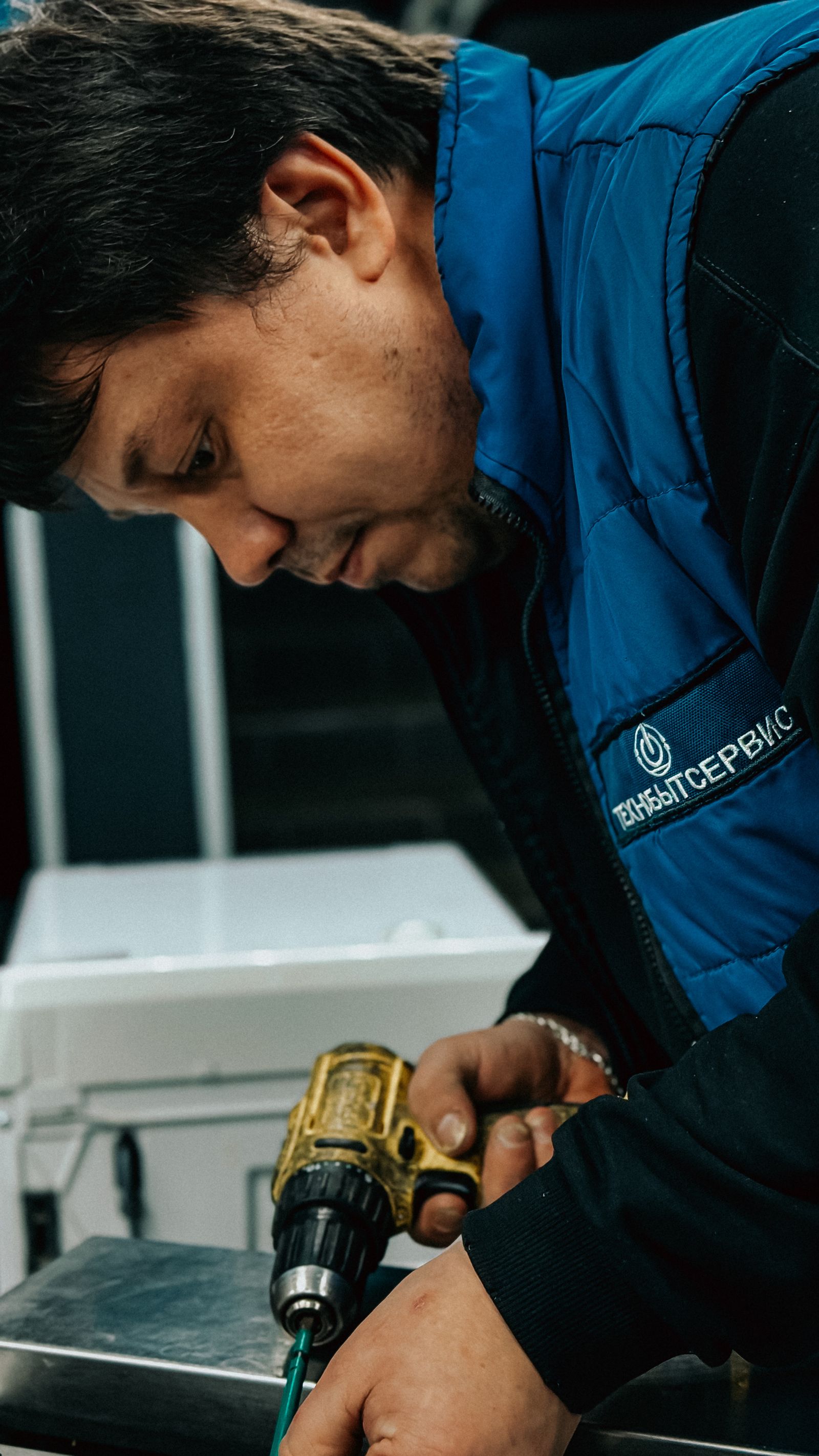 Close-up of a dark-haired technician in a blue vest bent over a workbench, driving a screw with a yellow cordless drill, concentrating on the fix.