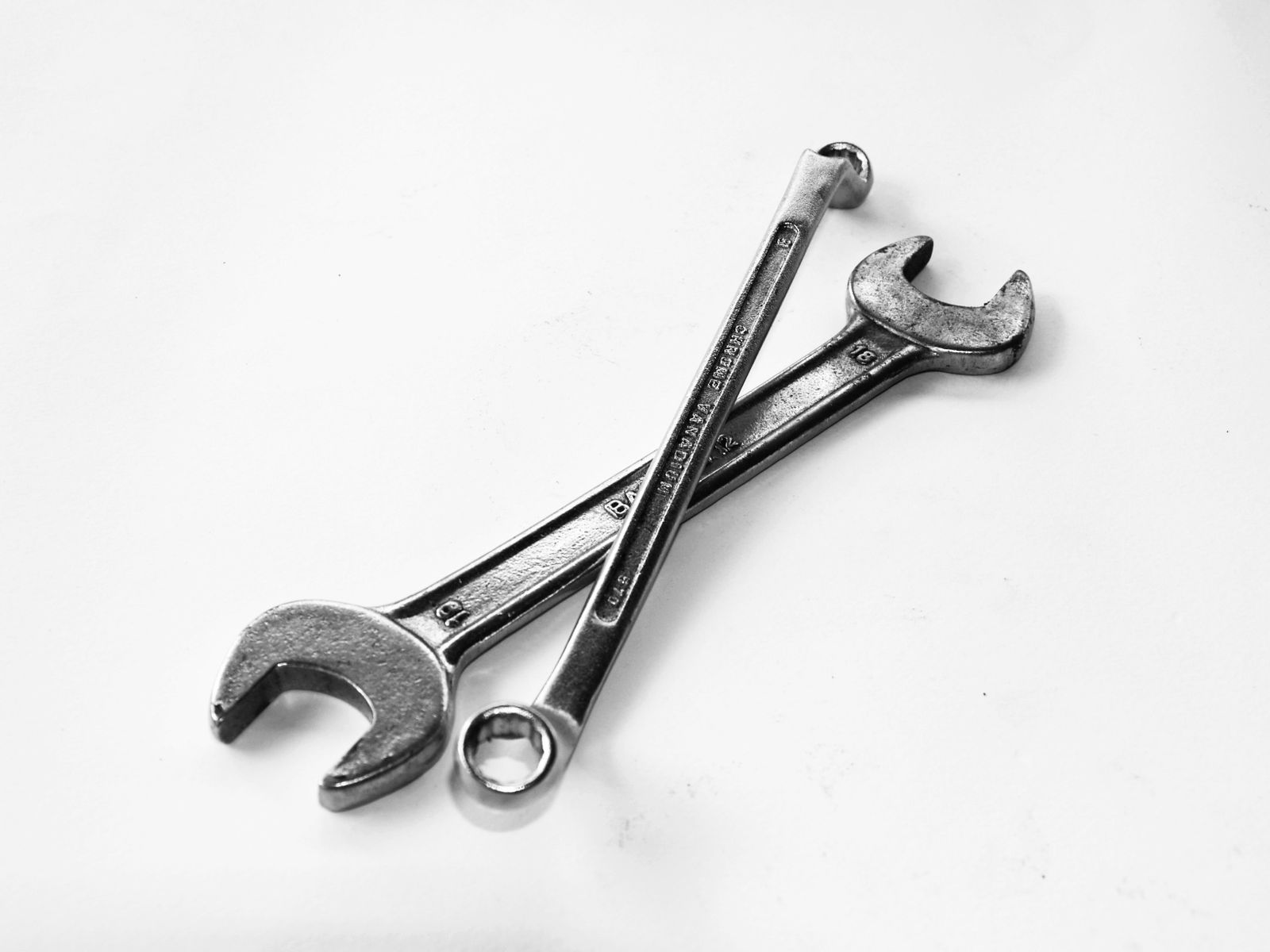 Two polished chrome combination wrenches crossed on a bright white background, desaturated studio shot.