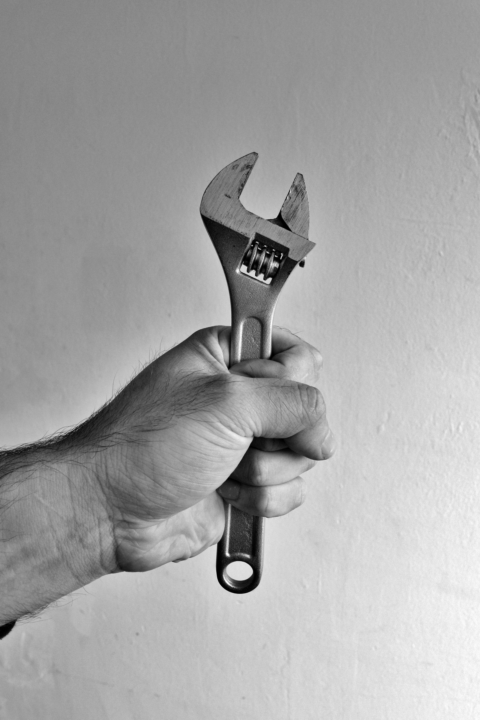 Black-and-white close-up of a man's hand and forearm gripping an adjustable wrench held upright against a plain light wall.