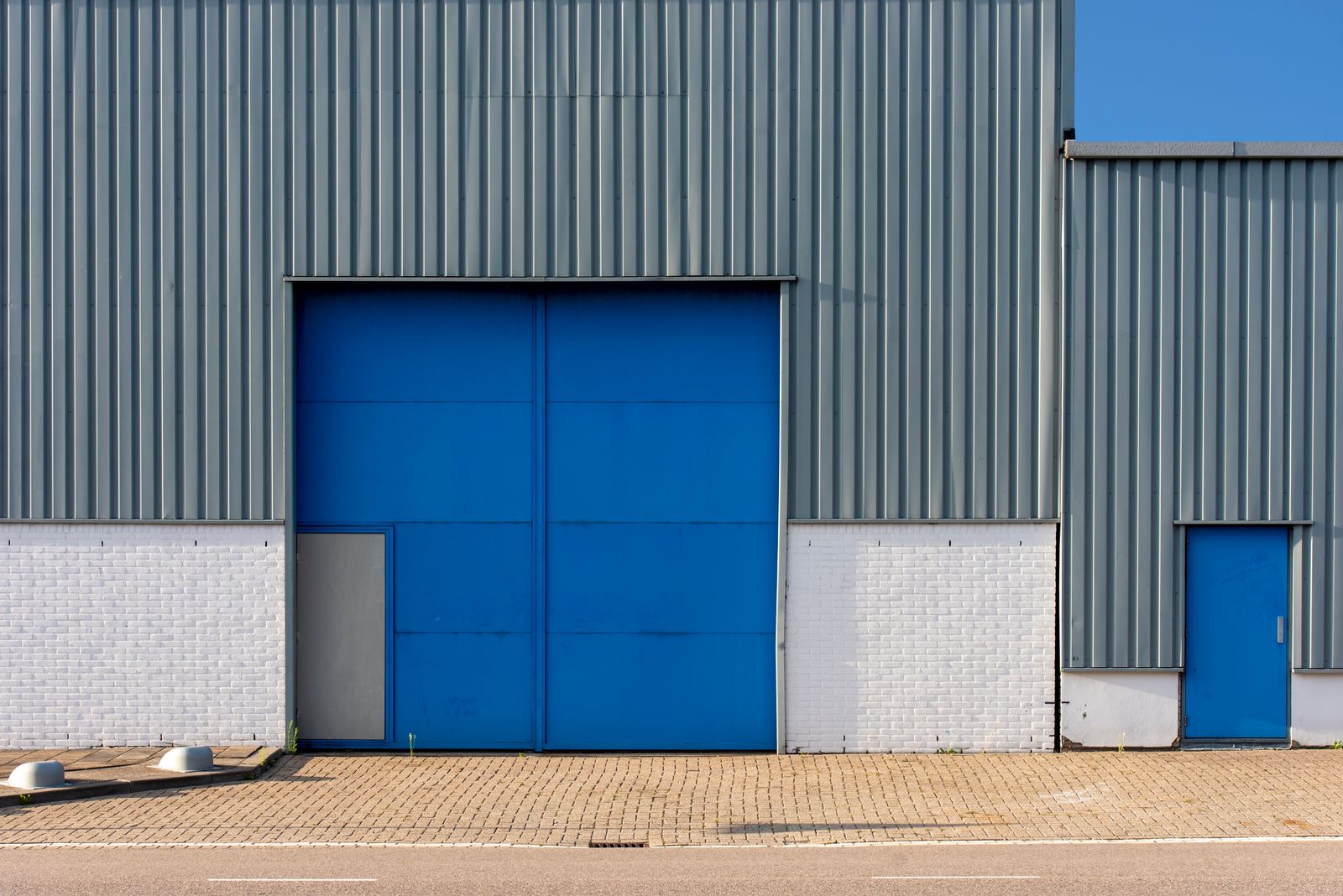 Industrial warehouse exterior with grey vertical metal siding, white brick base, and a tall blue double overhead sectional door (plus a smaller blue man-door at right) under a clear blue sky,…