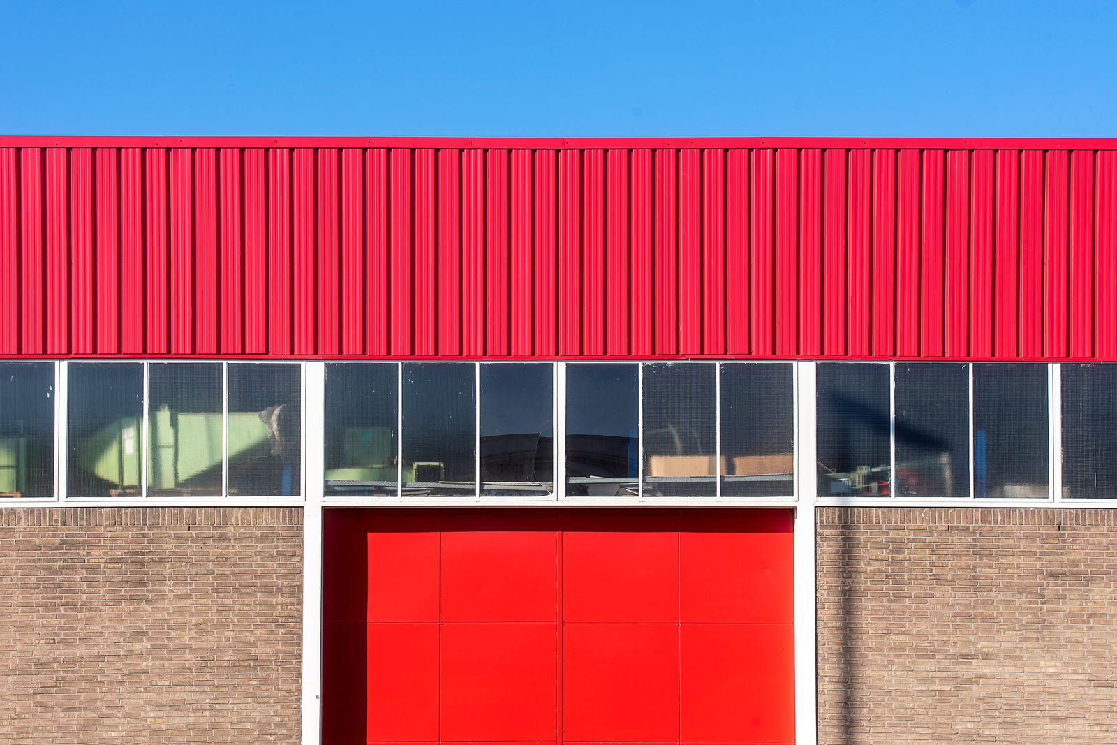 Commercial building facade with brown brick base, a band of clerestory windows, a red metal overhead door in the center, and red corrugated cladding above, under a clear blue sky.