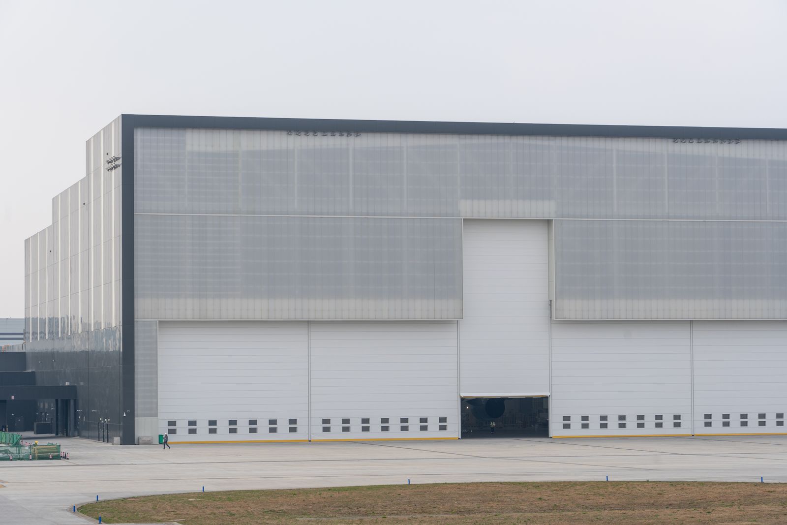 Large modern aircraft-hangar-style commercial building with translucent panel upper facade and a row of white industrial sectional/roller doors at ground level, expansive concrete apron in front…