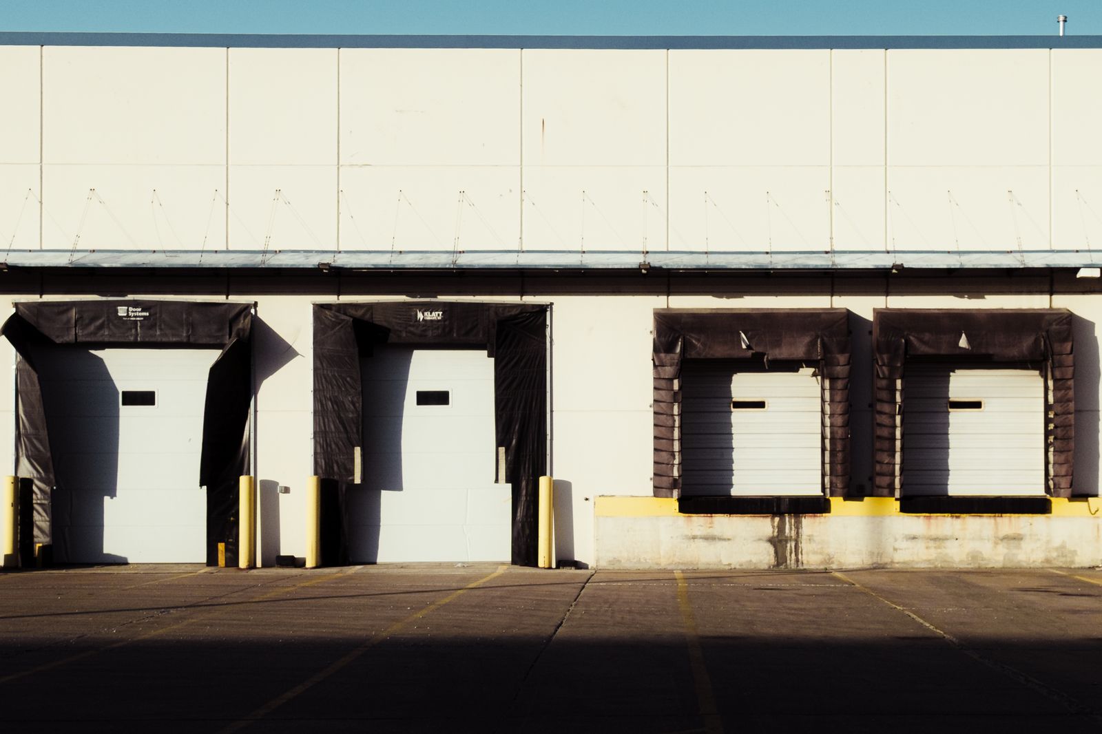 Warehouse loading dock exterior in warm low sunlight: four dock bays with black dock seals/shelters and white insulated dock doors above a concrete dock wall with yellow bumper posts, empty paved…